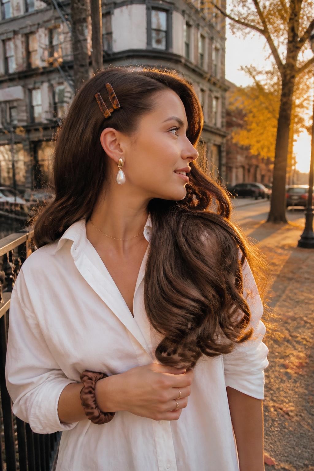 Photo of a woman wearing long glossy curls with a dramatic deep side part, one side pinned behind the ear using three amber barrettes, candid side-profile view on a softly blurred city street with golden leaves, warm sunset glow, statement earring visible on the pinned side and a silk scrunchie around her wrist, realistic curl movement in the breeze, latest iPhone photo quality.