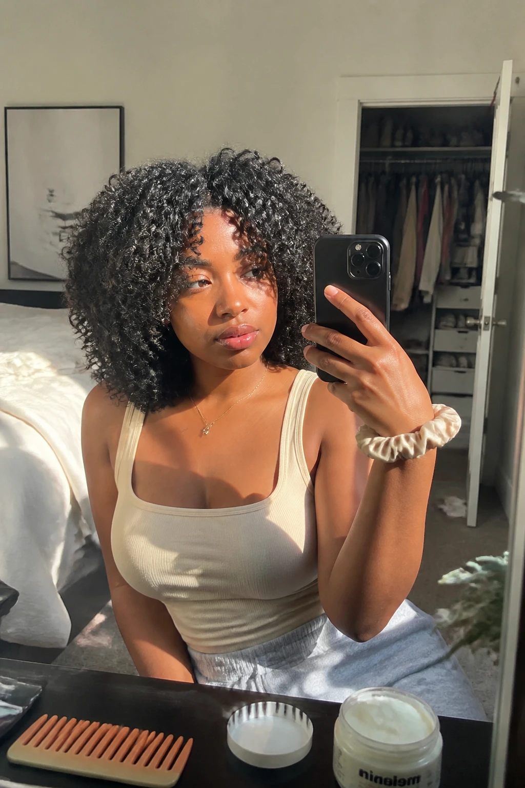 Image prompt

Photo of a Black woman wearing shoulder-length 4A curls in a softly rounded cut with dense defined coils, light face-framing layers beginning near the cheekbones, and balanced volume around the crown, candid mirror selfie in a sun-drenched bedroom, natural window light, containing a small lived-in detail such as a curl cream jar and wide-tooth comb on the dresser, a silk scrunchie on her wrist, and a slightly open closet behind her, realistic getting-ready moment, latest iPhone photo quality.