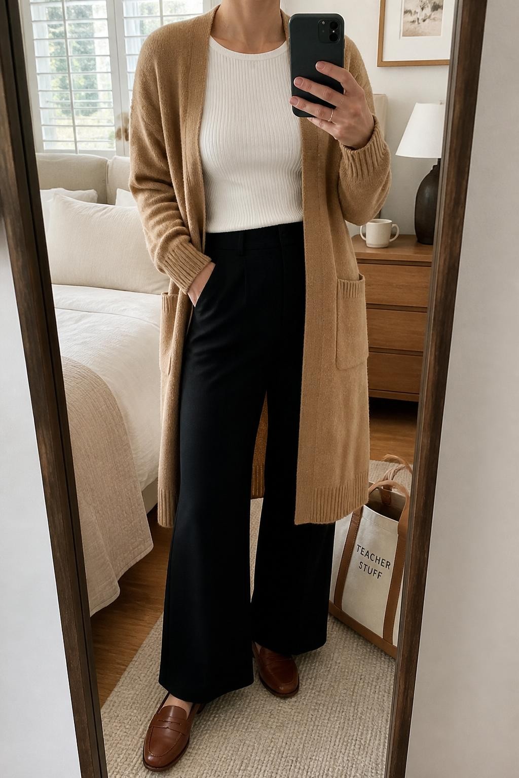 mirror selfie of a woman wearing a camel long cardigan over a white ribbed knit top, black wide-leg pants, brown leather loafers, and a tan canvas teacher tote, clearly showing the same clothing items, colors, fabrics, and accessories listed in the Outfit Pieces section, holding a phone in front of a mirror, cropped at NECK so the face does not show, with a relaxed casual selfie pose, in a cozy bedroom mirror setup, natural window light, including a tote bag resting on the floor and a coffee mug on a bedside table, latest iPhone photo quality.