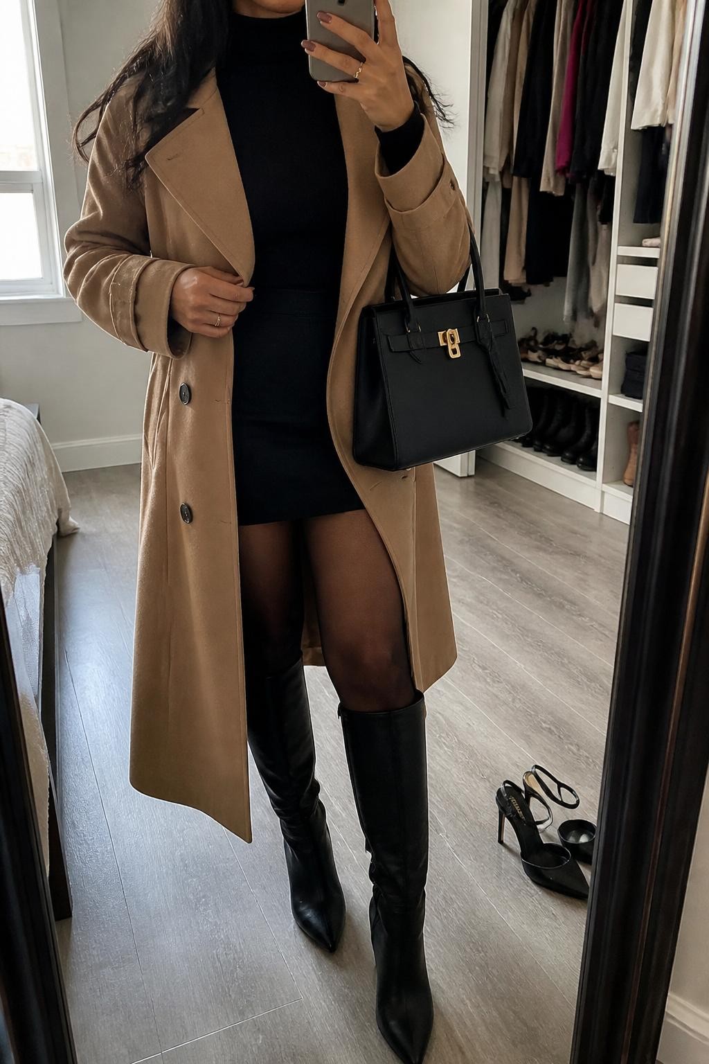 mirror selfie of an olive-toned Latina woman wearing a camel trench coat, black fitted turtleneck, black mini skirt, sheer black tights, black knee-high boots, and a structured black handbag, holding a phone in front of a mirror, cropped at NECK so the face does not show, relaxed casual selfie pose, in a walk-in closet mirror with natural window light, including an open closet with clothes visible and a pair of heels on the floor nearby, realistic latest iPhone photo quality.