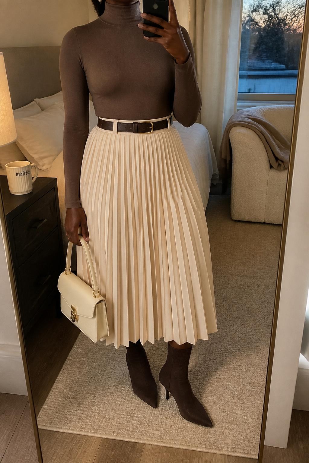 mirror selfie of a Black woman wearing the exact outfit described above, clearly showing dark brown sock boots with a slim heel, a cream pleated midi skirt, a mocha fitted turtleneck, a dark brown leather belt, a cream top-handle bag, and pearl earrings, holding a phone in front of a mirror, cropped at NECK so the face does not show, with a relaxed casual selfie pose, in a cozy bedroom mirror setup, evening golden hour, with a coffee mug on a bedside table and a folded blanket on a chair, latest iPhone photo quality.