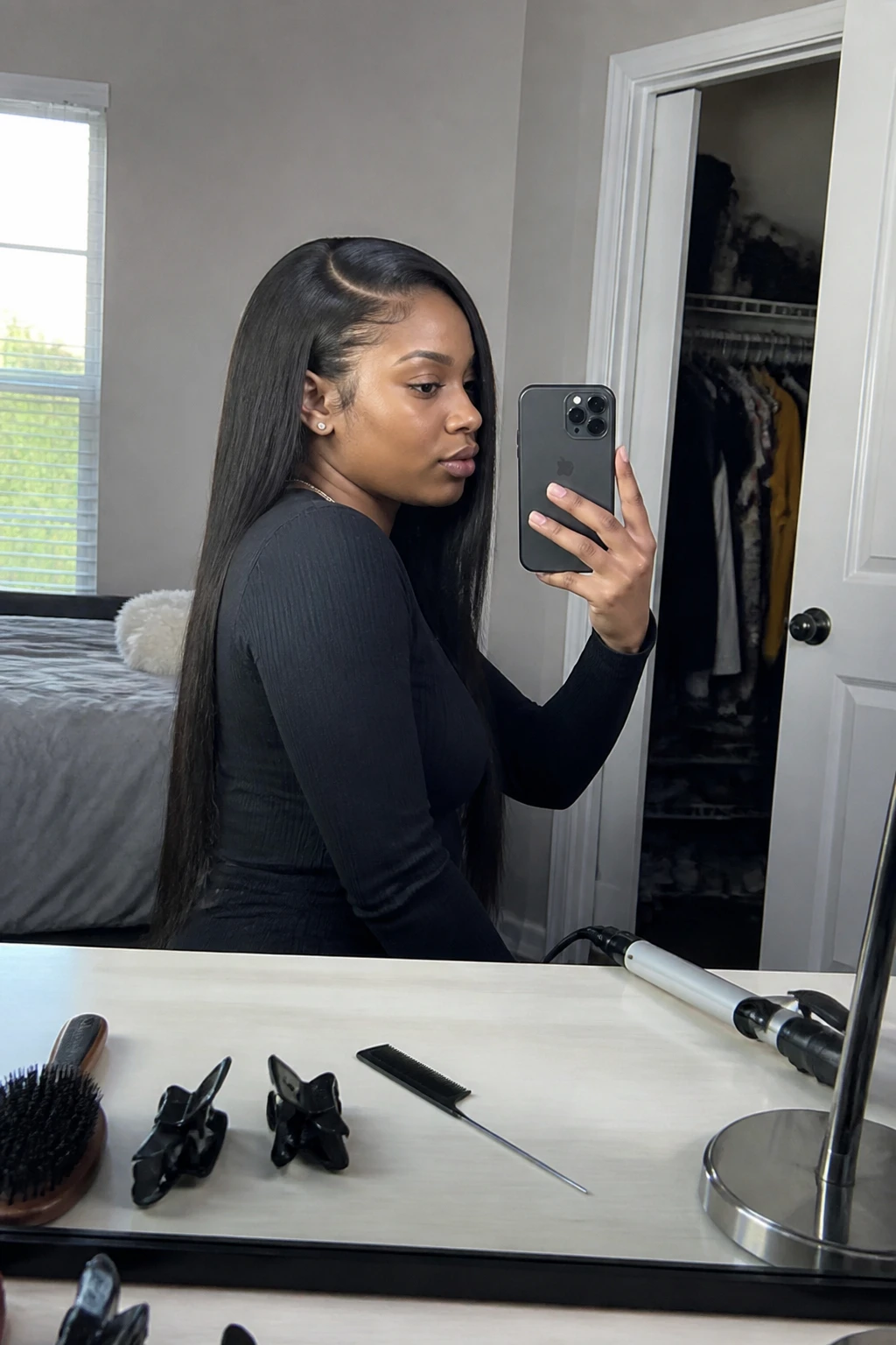 Image prompt

Photo of a woman wearing a long straight weave with smooth lengths, a clean side part, and subtle hidden volume at the crown, three-quarter side view captured in a bedroom mirror, natural window light, containing a small lived-in detail such as sectioning clips and a fine-tooth comb on the dresser, a curling iron nearby, and a slightly open closet behind her, realistic getting-ready moment, latest iPhone photo quality.