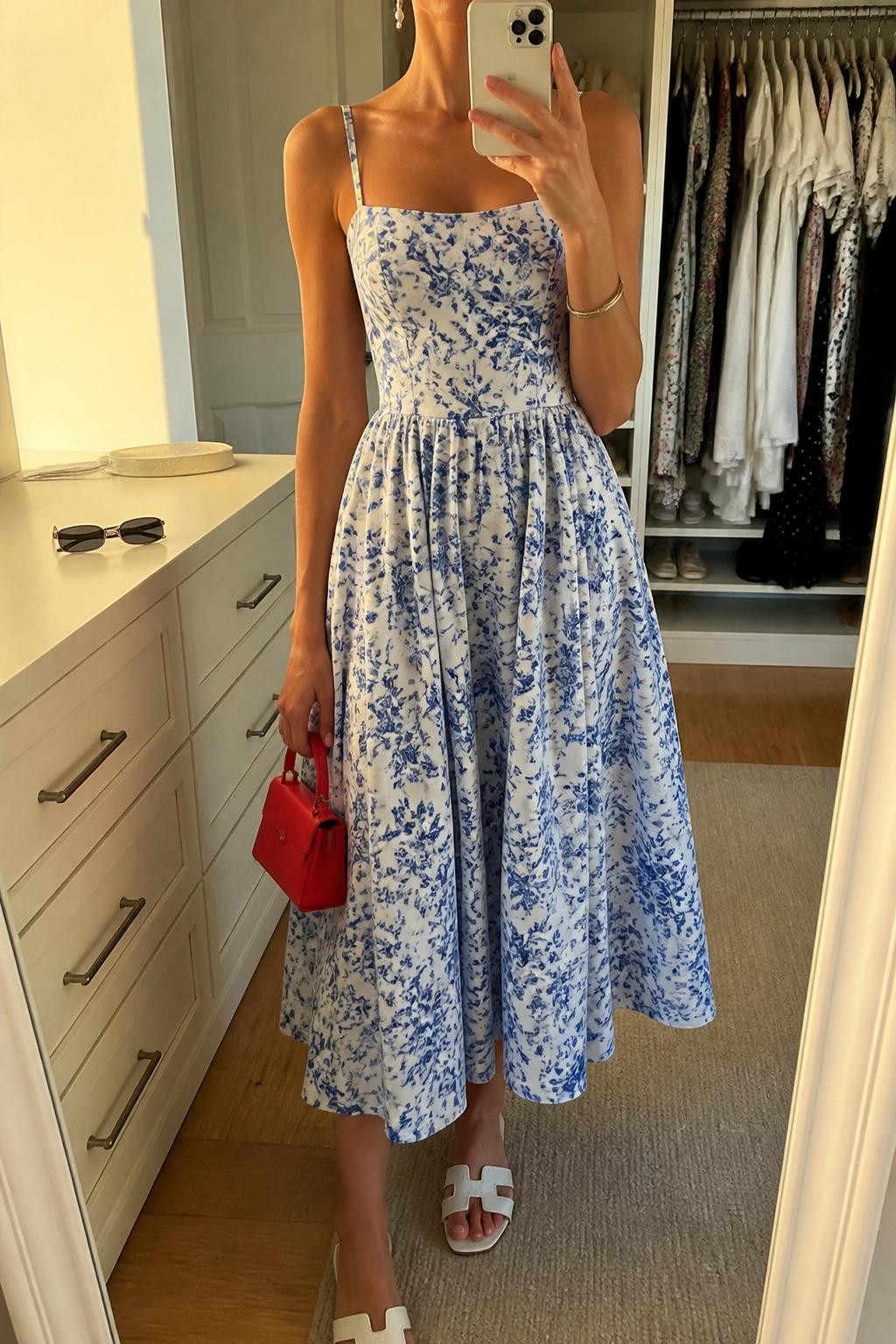 mirror selfie of a woman wearing a blue and white floral cotton sundress with thin straps and a flowing midi skirt, white flat sandals, red mini handbag, pearl drop earrings, and a slim silver bracelet, clearly showing the same clothing items, colors, fabrics, and accessories listed in the Outfit Pieces section, holding a phone in front of a mirror, cropped at NECK so the face does not show, with a relaxed casual selfie pose, in a walk-in closet mirror, evening golden hour, including an open closet with clothes visible and sunglasses placed on a dresser, latest iPhone photo quality.
