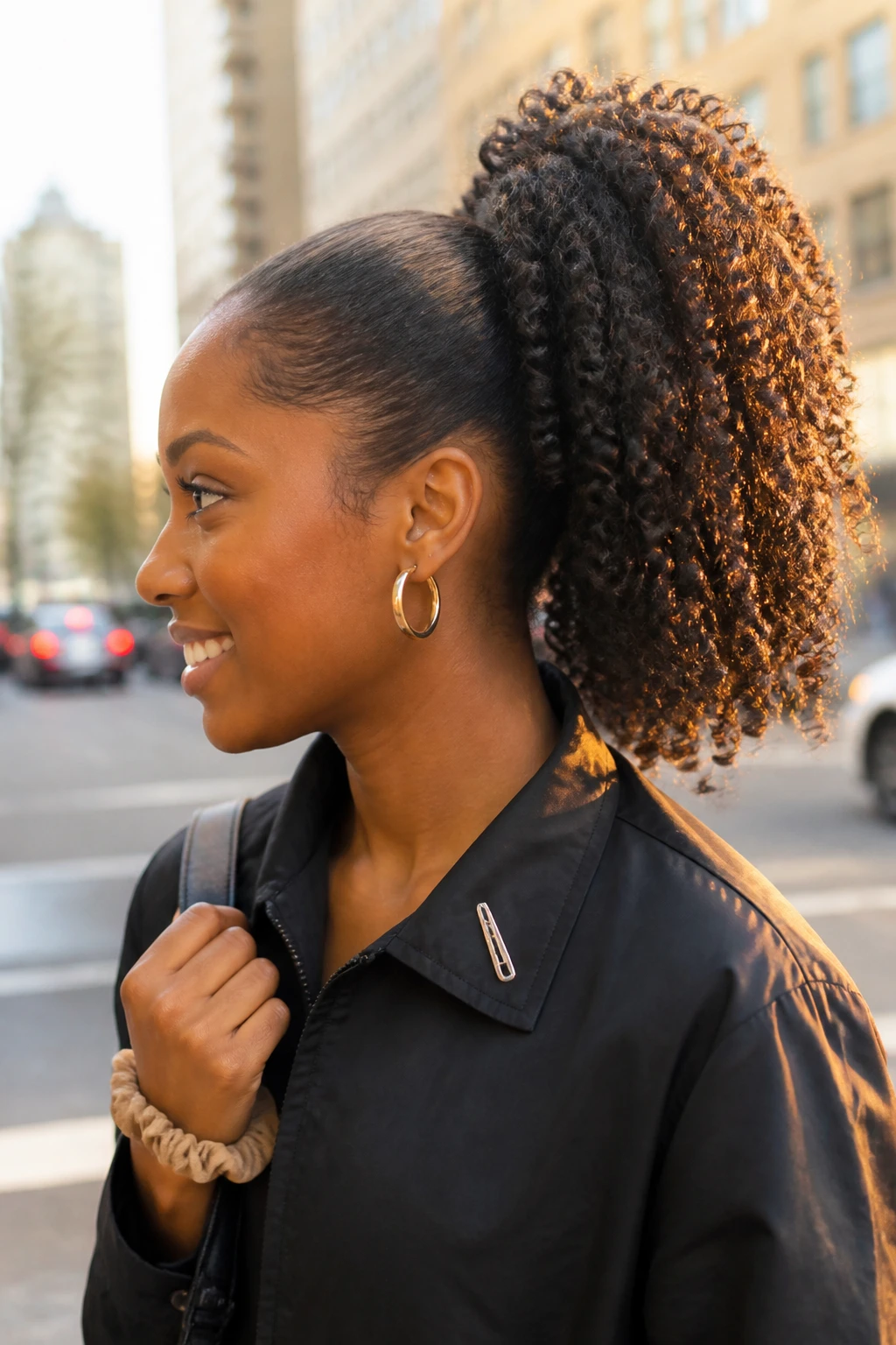 Image prompt
Photo of a Black woman wearing a high 3B curly ponytail with a smooth base, neatly wrapped hair section around the elastic, and full springy curls cascading from the crown, side profile view against a softly blurred city street background, golden hour glow, containing a small lived-in detail such as gold hoop earrings, a spare scrunchie on her wrist, and a slim hair clip attached to her jacket collar, realistic candid photograph, latest iPhone photo quality