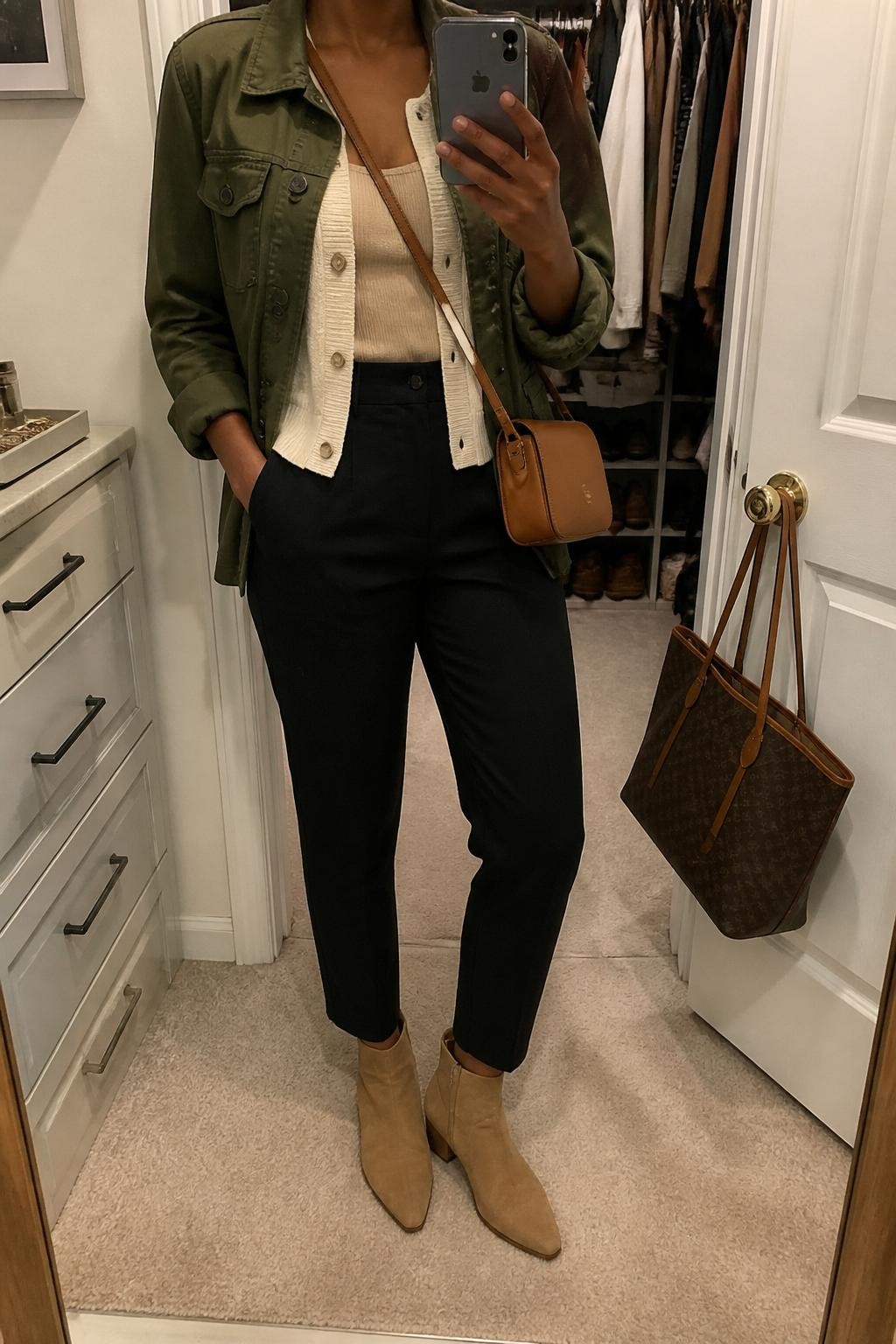 Mirror selfie of a Black woman with deep brown skin wearing an olive green cotton utility jacket over a lightweight cream knit cardigan and a fitted beige ribbed tank top, black high-waisted ankle trousers, tan suede ankle boots, and a compact cognac leather crossbody bag, clearly showing the exact clothing items, colors, fabrics, and accessories listed, holding a latest-model iPhone in front of a mirror, cropped at the neck so the face does not show, relaxed casual selfie pose, in a walk-in closet mirror with soft indoor lighting, an open closet with clothes visible and a handbag hanging on a door handle, realistic uncluttered setting, latest iPhone photo quality.