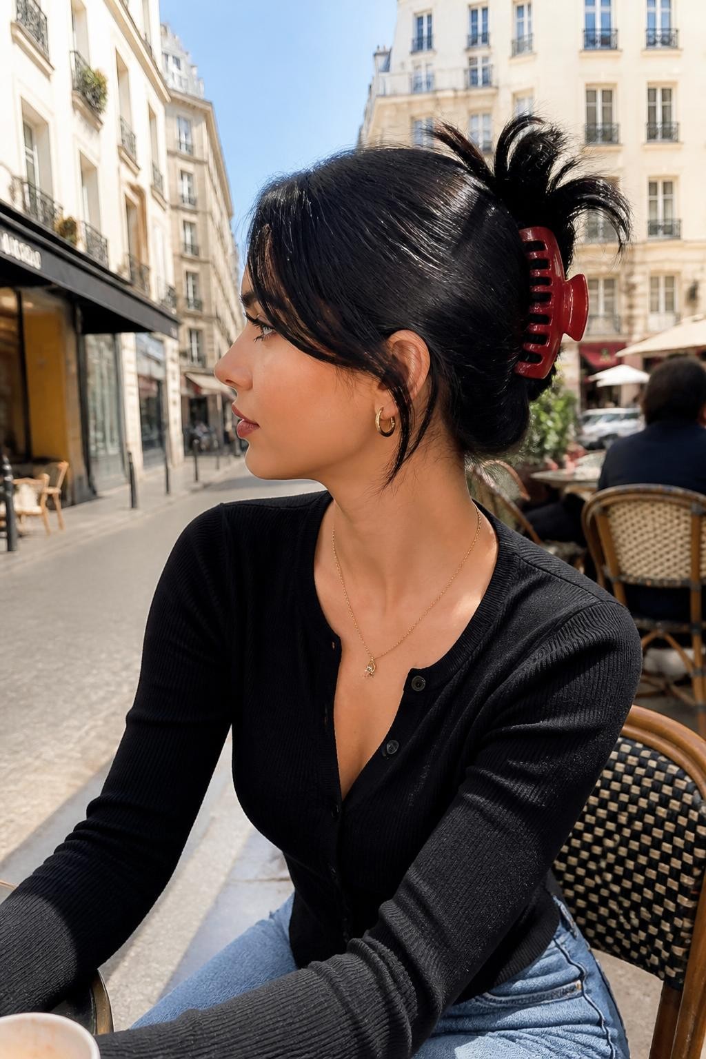 A realistic fashion beauty photo of a woman with long glossy black hair styled in a claw clip updo with flipped ends, hair twisted upward and clipped with a large red claw clip, ends fanning out at the top with soft flipped shape, sleek side bangs, fitted cardigan, retro-inspired makeup, bright city café background, trendy 90s hairstyle inspiration, no text, no watermark.