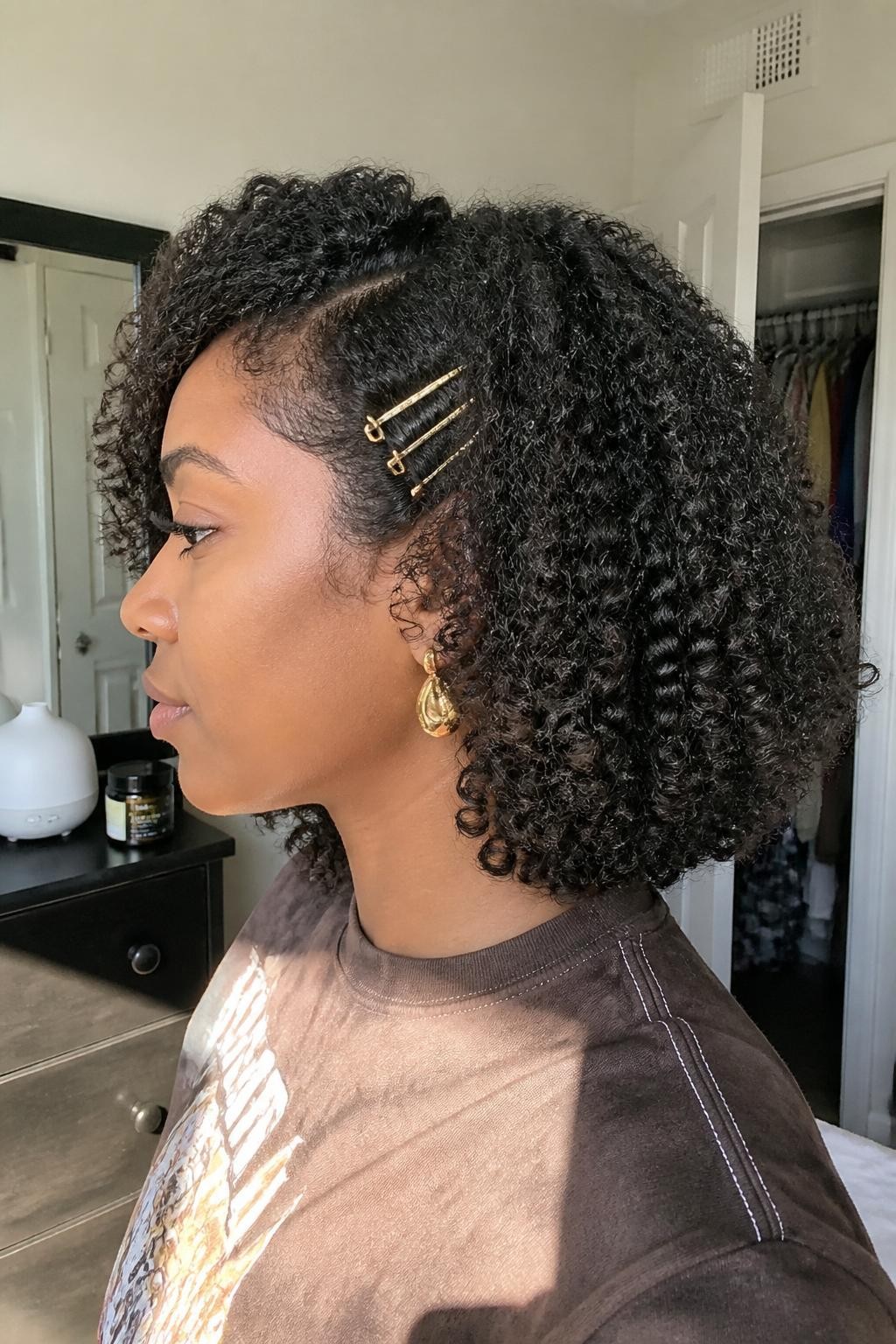 Photo of a Black woman wearing a shoulder-grazing curly bob with defined ringlets, rounded volume, and three slim gold clips placed above one ear, side-profile view in a sun-drenched bedroom, natural window light, diffuser and curl gel resting on the dresser, statement earring peeking below the clips and a slightly open closet behind her, realistic curl shrinkage and soft frizz, latest iPhone photo quality.