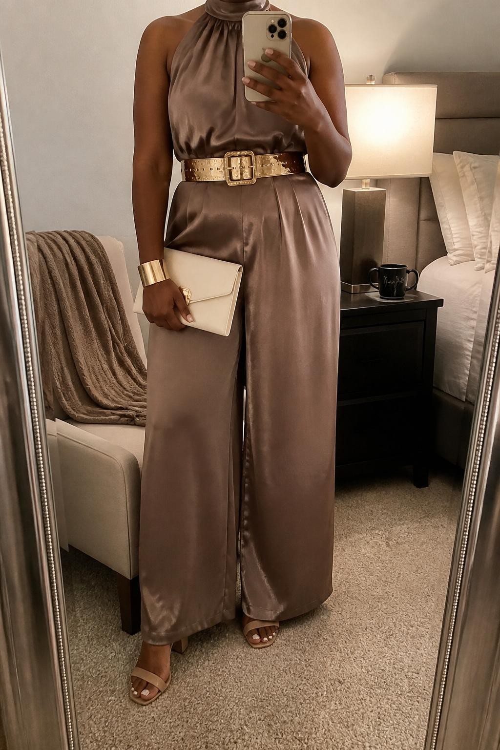 mirror selfie of a Black woman wearing the exact outfit described above, clearly showing a mocha satin wide-leg jumpsuit, a gold statement belt at the waist, nude block heels, a cream clutch, and gold cuff bracelet, holding a phone in front of a mirror, cropped at NECK so the face does not show, with a relaxed casual selfie pose, in a cozy bedroom mirror setup, soft indoor lighting, with a folded blanket on a chair and a coffee mug on a bedside table, latest iPhone photo quality.