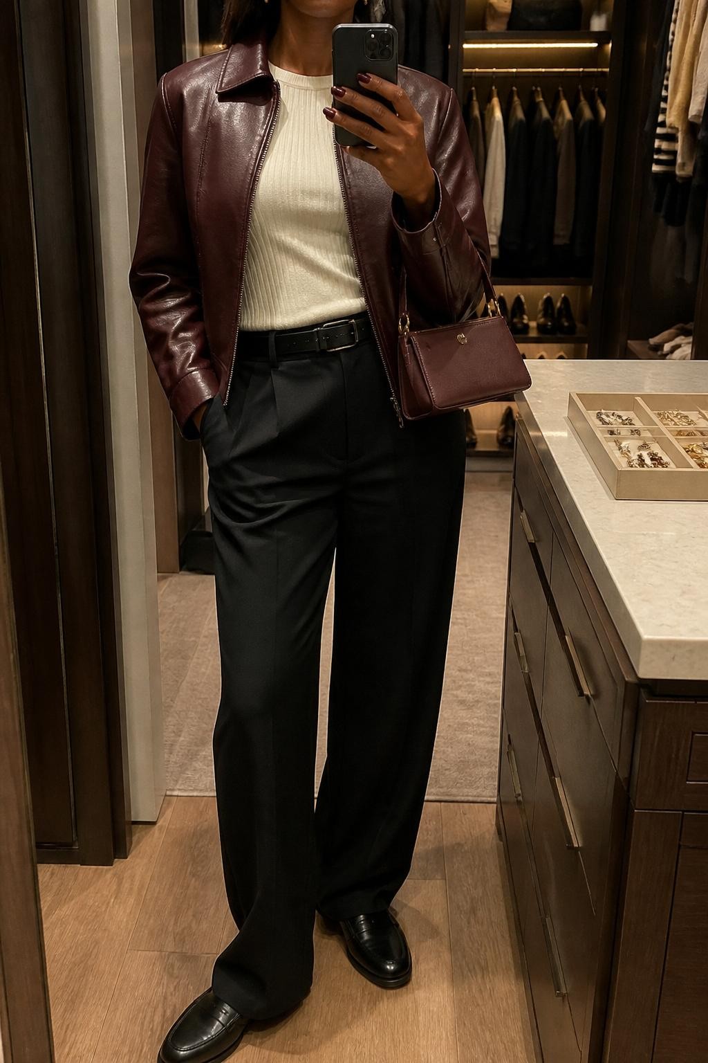 mirror selfie of a Black woman wearing the exact outfit described above, clearly showing a burgundy leather jacket layered over a cream ribbed knit top, black wide-leg trousers, black loafers, a burgundy mini handbag, and small gold stud earrings, holding a phone in front of a mirror, cropped at NECK so the face does not show, with a relaxed casual selfie pose, in a walk-in closet mirror, warm moody lighting, with an open closet with clothes visible and a jewelry tray on the vanity, latest iPhone photo quality.