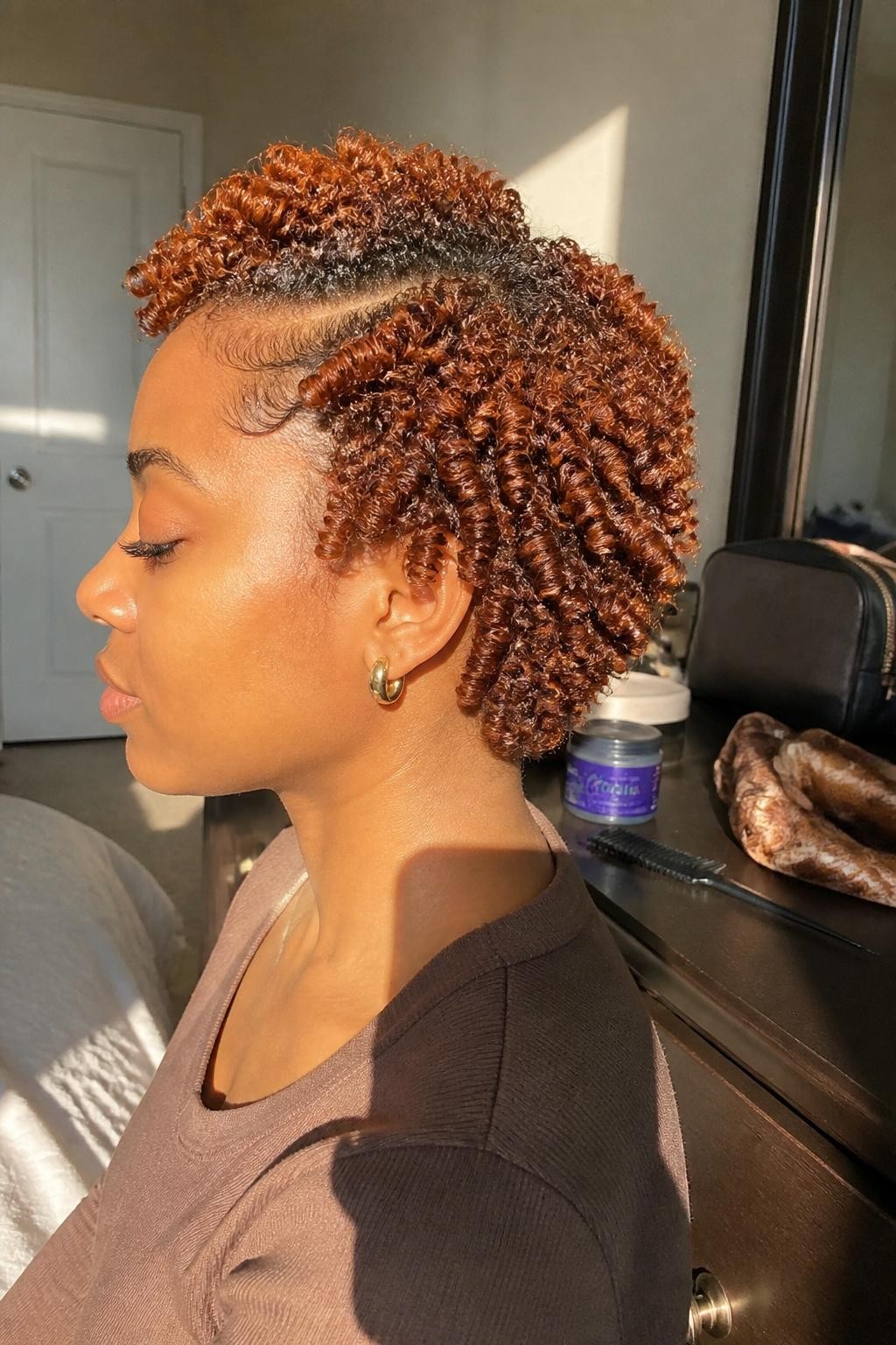 Photo of a Black woman wearing short copper finger coils with a deep side part, defined spiral texture, and softly shaped edges, side-profile view in a sun-drenched bedroom, warm natural window light, curl gel and a rat-tail comb on the dresser, small gold earrings visible and a silk scarf resting beside a makeup bag, realistic coil definition and natural texture, latest iPhone photo quality.