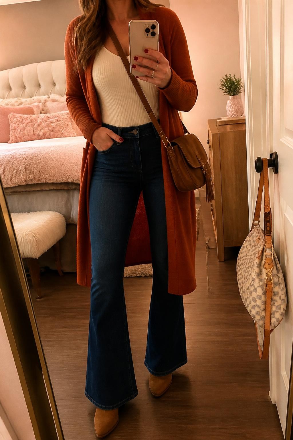 mirror selfie of a woman wearing the exact outfit described above, clearly showing a burnt orange long coatigan, cream ribbed bodysuit, dark blue flare jeans, tan heeled boots, and a brown suede crossbody bag, holding a phone in front of a mirror, cropped at NECK so the face does not show, with a relaxed casual selfie pose, in a feminine-themed bedroom, warm moody lighting, with a handbag hanging on a door handle and a small plant on a dresser, latest iPhone photo quality.