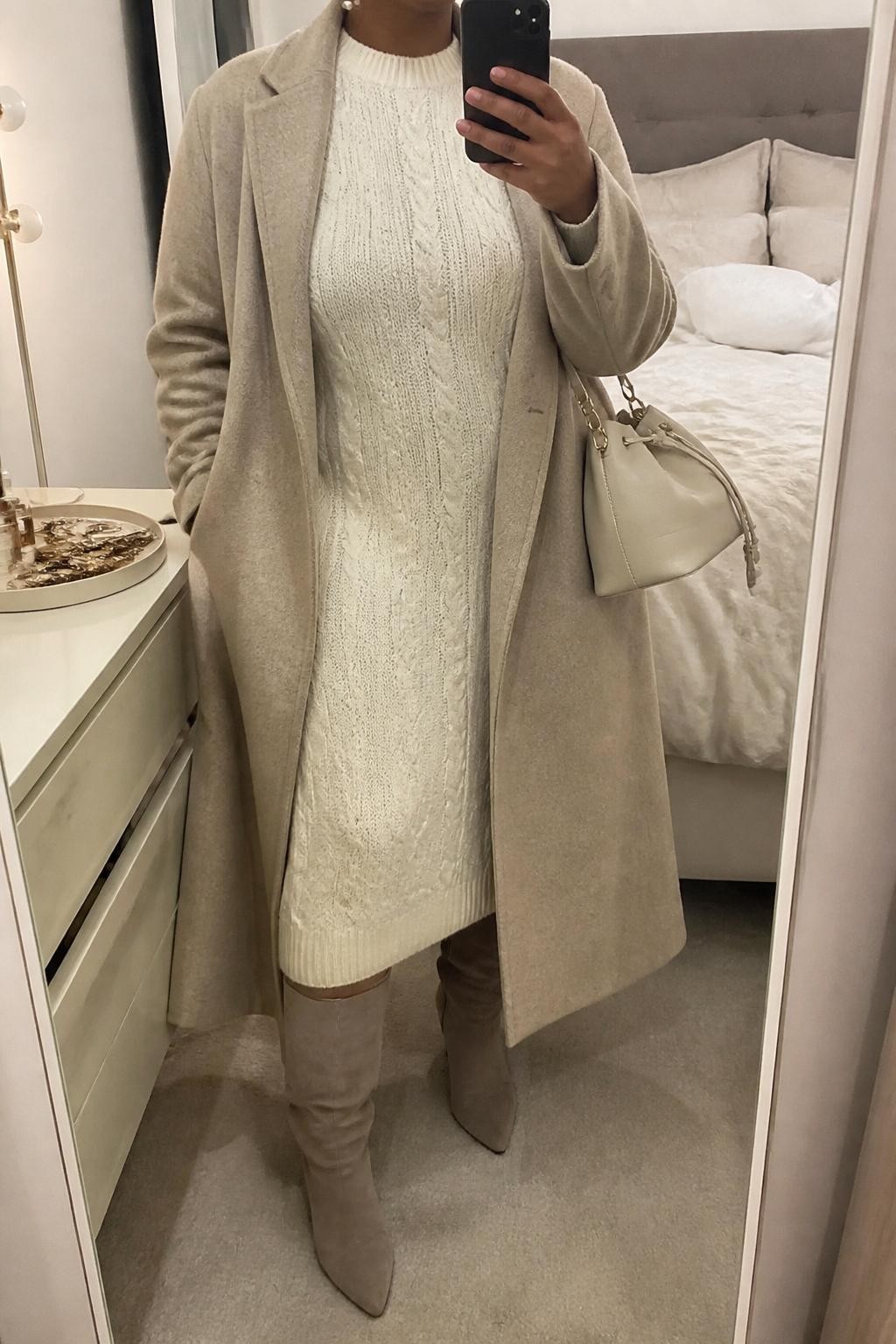 mirror selfie of a plus size Black woman wearing the exact outfit described above, an oatmeal relaxed midi cable knit dress, taupe suede wide-calf tall boots, soft beige long wool coat, cream leather mini bucket bag, and pearl stud earrings, clearly showing the same clothing items, colors, fabrics, and accessories listed in the Outfit Pieces section, holding a phone in front of a mirror, cropped at NECK so the face does not show, with a relaxed casual selfie pose, in a cozy bedroom mirror setup, soft indoor lighting, including soft pillows on an unmade bed and a jewelry tray on the vanity, latest iPhone photo quality.
