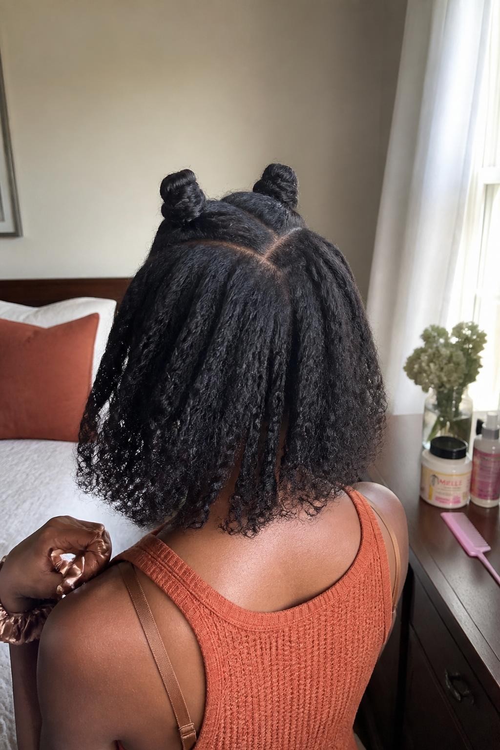Photo of a Black woman wearing a shoulder-length braid-out lob with defined textured waves and two small Bantu knots positioned near the crown, back-of-head view in a softly lit bedroom, warm window light, curl cream and small sectioning comb on the dresser, silk scrunchie around her wrist and a rust knit top visible, realistic natural texture and soft flyaways, latest iPhone photo quality.