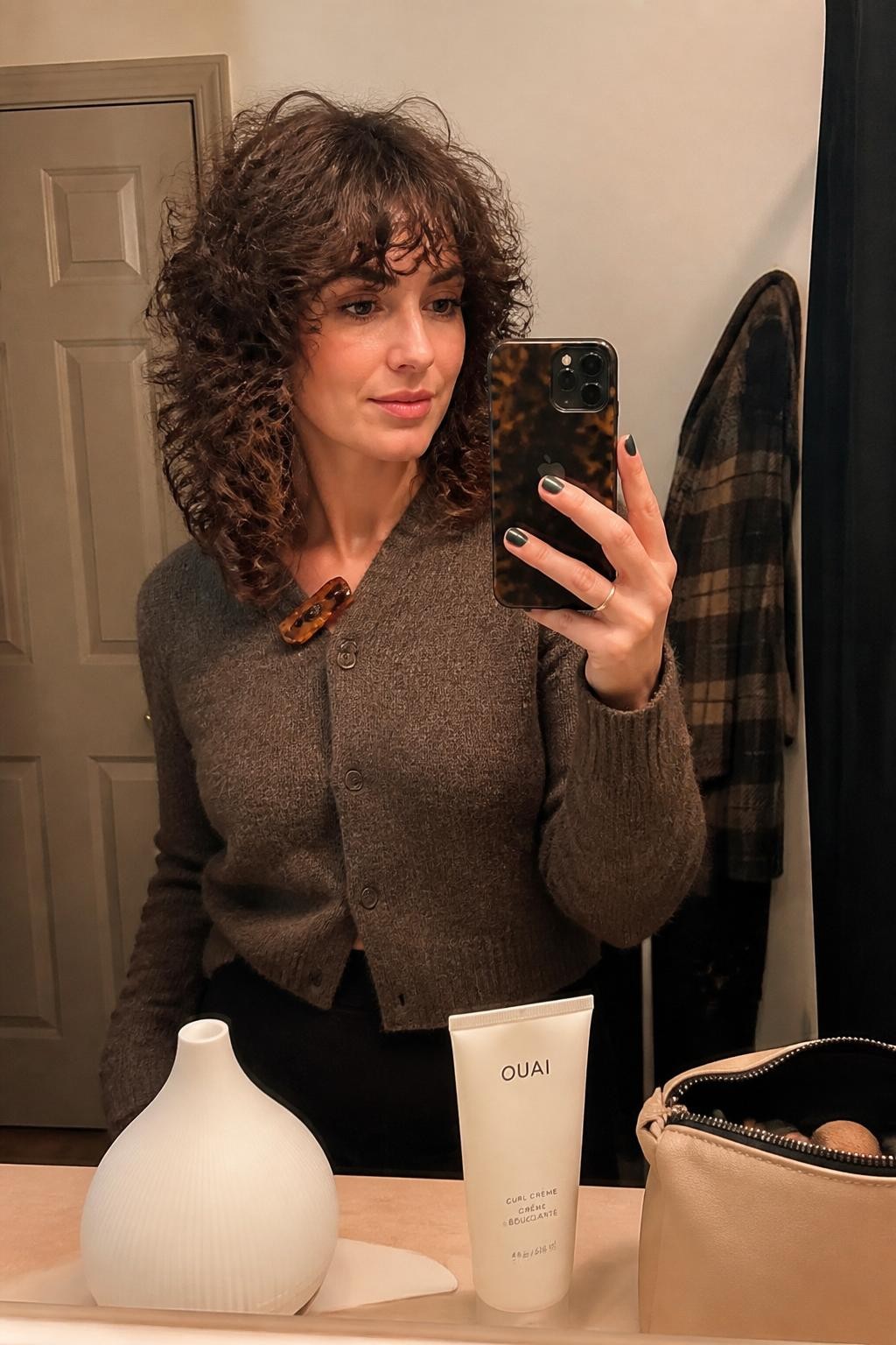 Photo of a woman wearing a shoulder-length curly shag with short airy crown layers, bottleneck bangs, and softly tapered ends, candid mirror selfie in a warm bathroom vanity mirror, soft indoor lighting, diffuser and curl cream resting on the counter beside a makeup pouch, tortoiseshell clip attached to her cardigan collar and a plaid coat visible behind her, realistic curl separation and flyaways, latest iPhone photo quality.