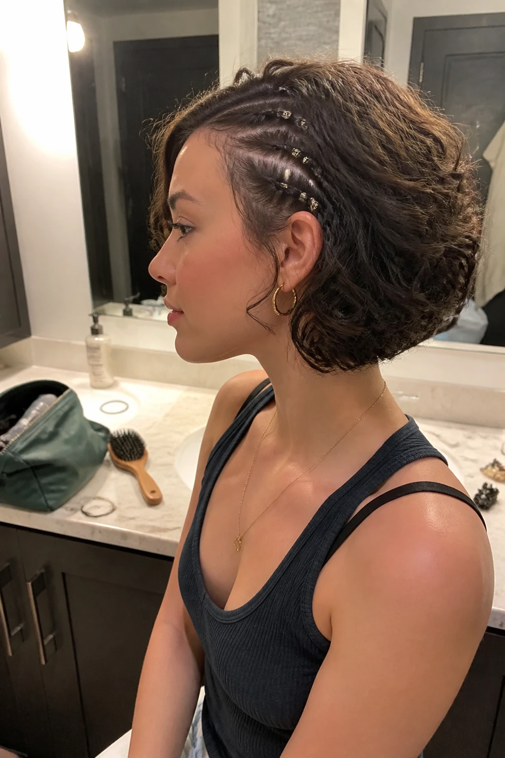 Image prompt

Photo of a woman wearing a short curly bob with two thin side braids near one temple, loose defined curls, and small silver cuffs at the braid ends, side profile view beside a bathroom vanity mirror, soft indoor lighting, containing a small lived-in detail such as tiny elastics on the counter, a makeup bag nearby, and a hairbrush resting beside the sink, realistic getting-ready moment, latest iPhone photo quality.