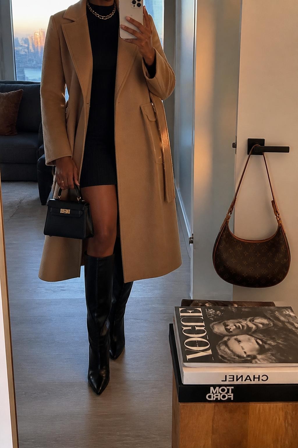 mirror selfie of a Black woman wearing the exact outfit described above, clearly showing a camel wrap coat, black fitted sweater dress, black knee-high boots, gold chain necklace, and a small black top-handle bag, holding a phone in front of a mirror, cropped at NECK so the face does not show, with a relaxed casual selfie pose, in a stylish city apartment, evening golden hour, with a handbag hanging on a door handle and a stack of fashion magazines on a side table, latest iPhone photo quality.