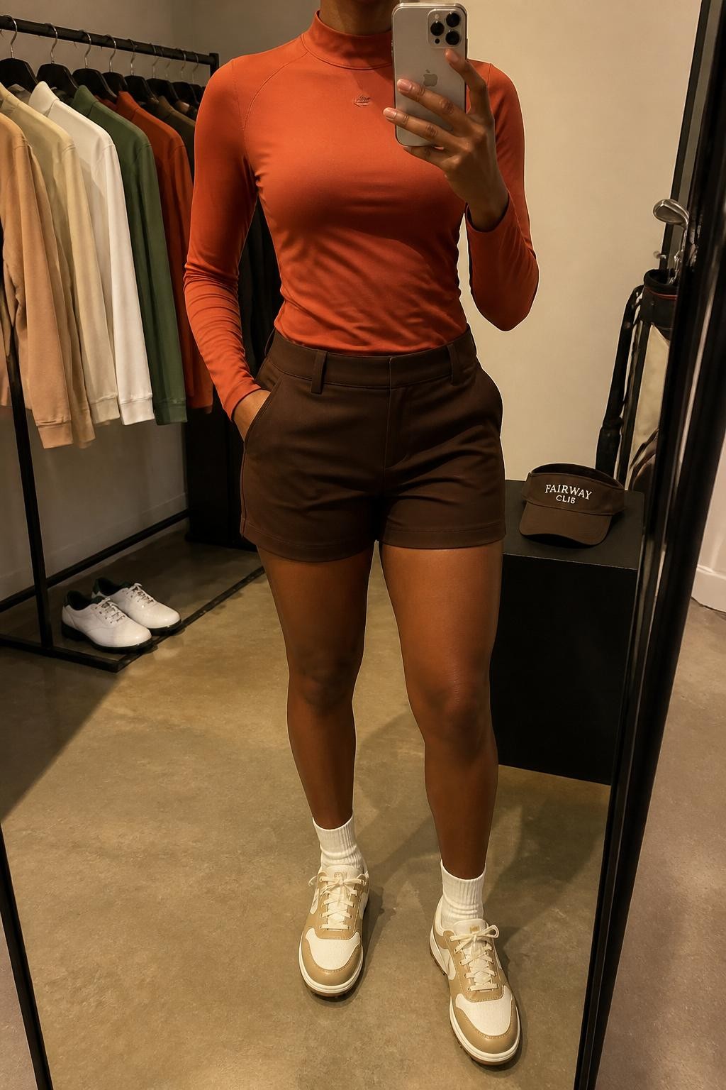 Mirror selfie of a Black woman with warm brown skin wearing a fitted rust-orange long-sleeve mock-neck performance top, chocolate brown tailored golf shorts with a mid-thigh length, cream ribbed ankle socks, tan-and-cream spikeless golf sneakers, and a chocolate brown visor, clearly showing the exact clothing items, colors, fabrics, and accessories listed, holding a latest-model iPhone in front of a mirror, cropped at the neck so the face does not show, relaxed casual selfie pose, in a fashion studio mirror corner with warm moody lighting, a clothing rack with golf tops behind and a pair of golf shoes on the floor, natural unstaged composition, latest iPhone photo quality.
