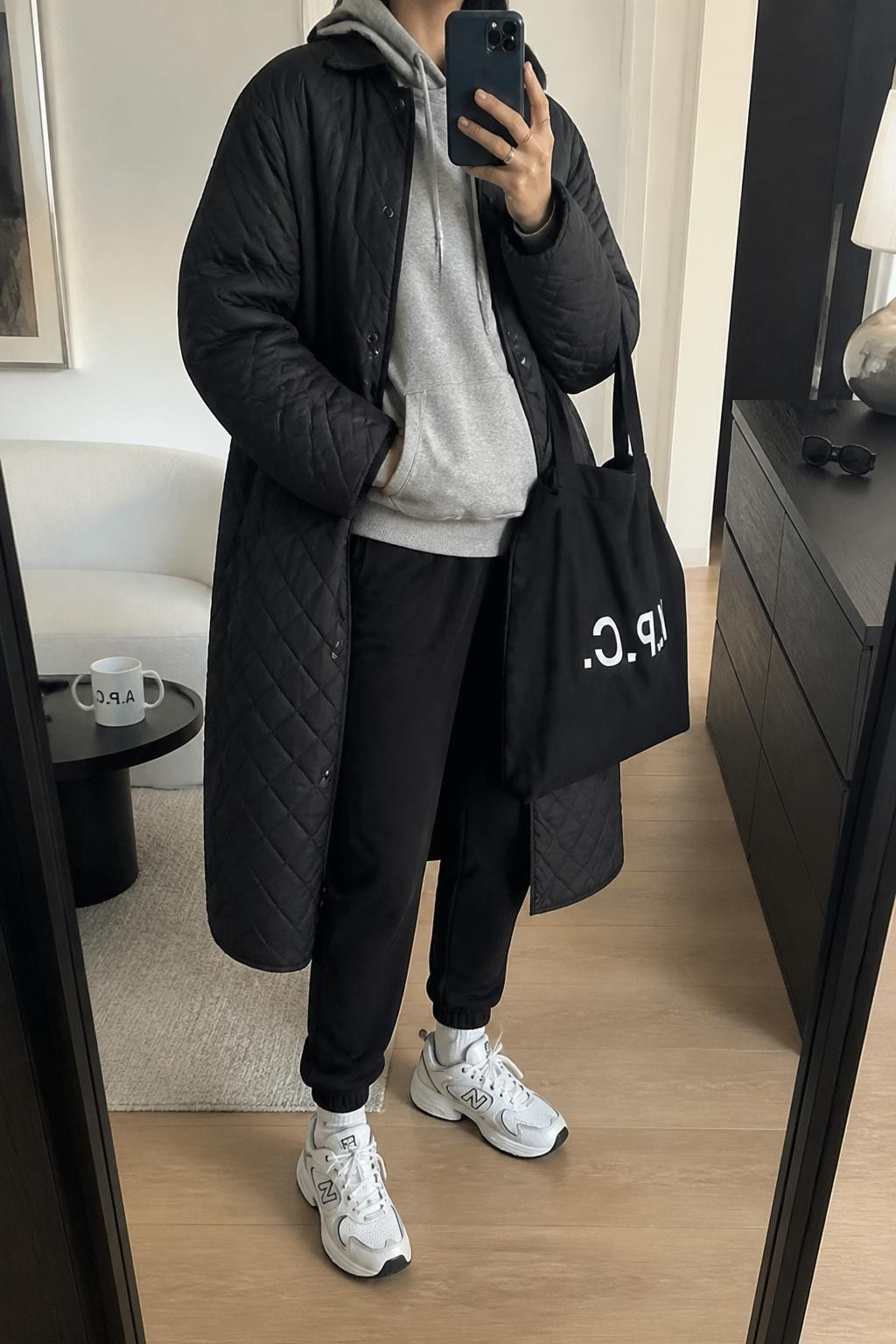 mirror selfie of a woman wearing a black quilted long coat over a gray hoodie, black jogger pants, white chunky sneakers, and a black canvas tote bag, clearly showing the same clothing items, colors, fabrics, and accessories listed in the Outfit Pieces section, holding a phone in front of a mirror, cropped at NECK so the face does not show, with a relaxed casual selfie pose, in a stylish city apartment, soft indoor lighting, including a coffee mug on a side table and sunglasses placed on a dresser, latest iPhone photo quality.