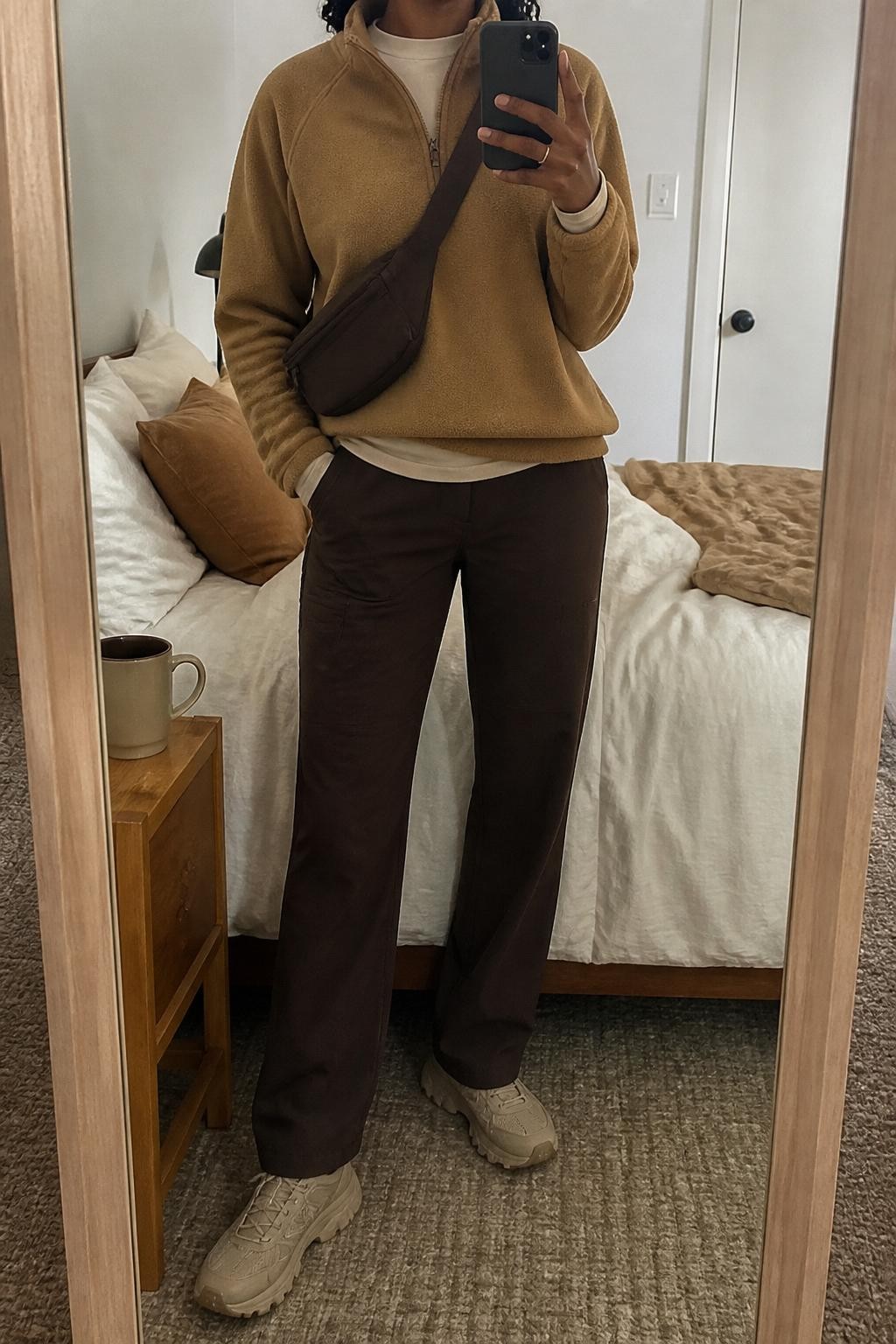 mirror selfie of a Black woman wearing the exact outfit described above, clearly showing a camel fleece half-zip pullover, chocolate brown straight-leg hiking pants, cream moisture-wicking base layer, beige trail sneakers, and a dark brown belt bag worn crossbody, holding a phone in front of a mirror, cropped at NECK so the face does not show, with a relaxed casual selfie pose, in a cozy bedroom mirror setup, soft indoor lighting, with a coffee mug on a bedside table and soft pillows on an unmade bed, latest iPhone photo quality.