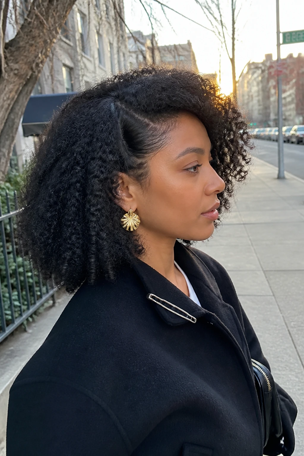 Image prompt

Photo of a Latina woman wearing stretched shoulder-length 4A curls with a deep side part, soft elongated coils, lifted roots, and full volume swept across one side, side profile view against a softly blurred city street background, golden hour glow, containing a small lived-in detail such as statement earrings, a slim hair clip attached to her jacket collar, and a small handbag near her elbow, realistic candid photograph, latest iPhone photo quality.