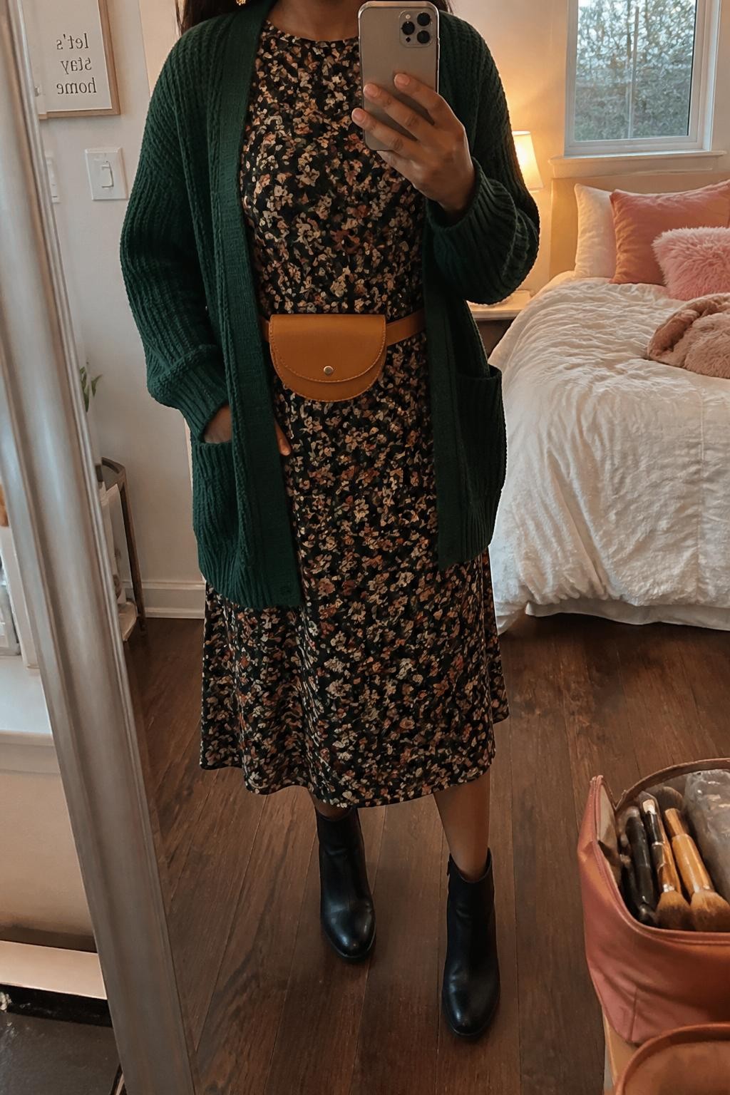 mirror selfie of a Black woman wearing the exact outfit described above, clearly showing a forest green chunky cardigan, black floral midi dress with rust and cream flowers, black ankle boots, tan belt bag, and small gold earrings, holding a phone in front of a mirror, cropped at NECK so the face does not show, with a relaxed casual selfie pose, in a feminine-themed bedroom, evening golden hour, with a makeup brush or cosmetic bag on the counter and soft pillows on an unmade bed, latest iPhone photo quality.