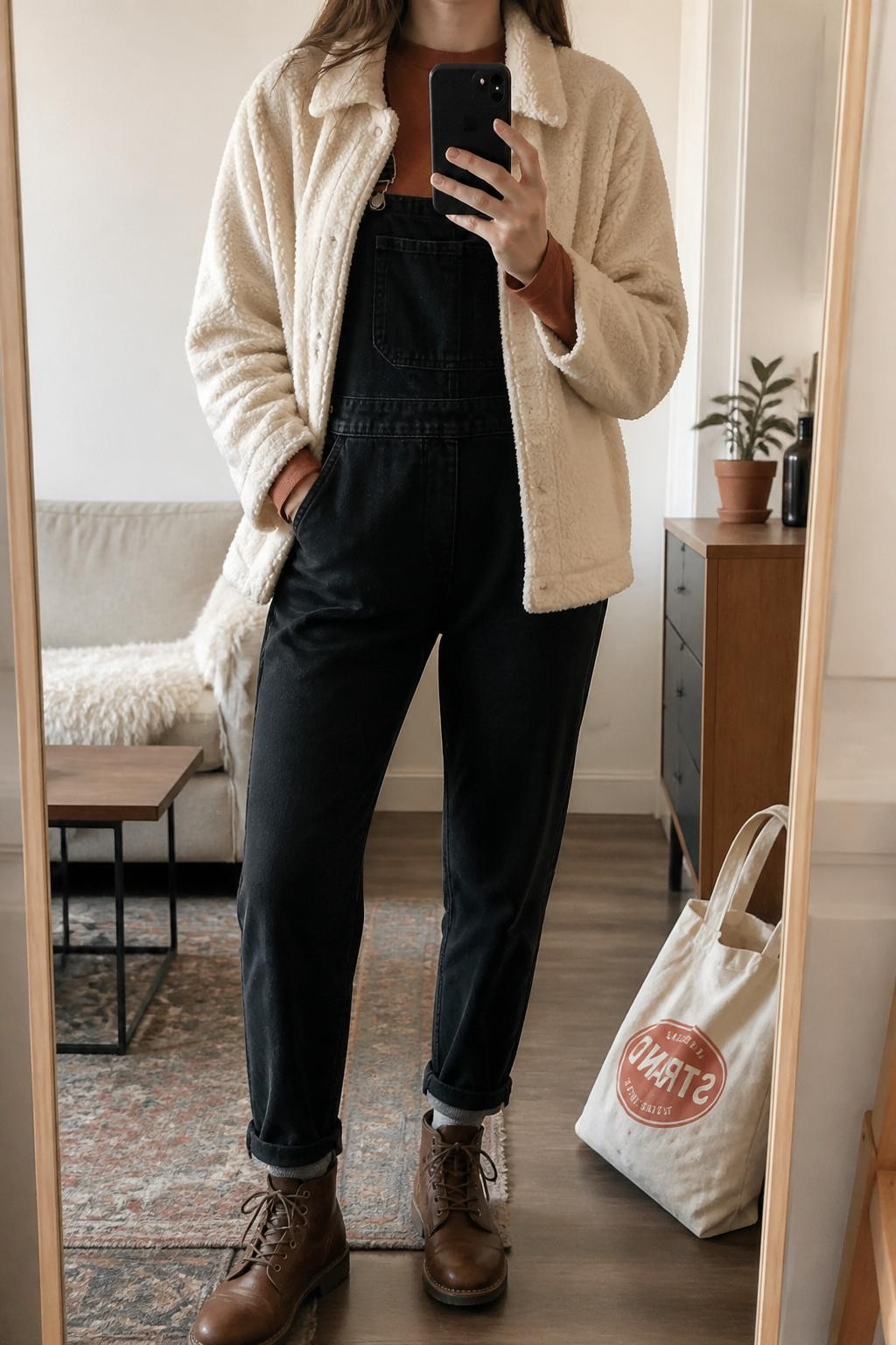 mirror selfie of a woman wearing the exact outfit described above, clearly showing a cream sherpa jacket, black denim overalls, rust long-sleeve tee, brown lace-up boots, and a canvas tote bag, holding a phone in front of a mirror, cropped at NECK so the face does not show, with a relaxed casual selfie pose, in a stylish city apartment, soft indoor lighting, with a tote bag resting on the floor and a small plant on a dresser, latest iPhone photo quality.