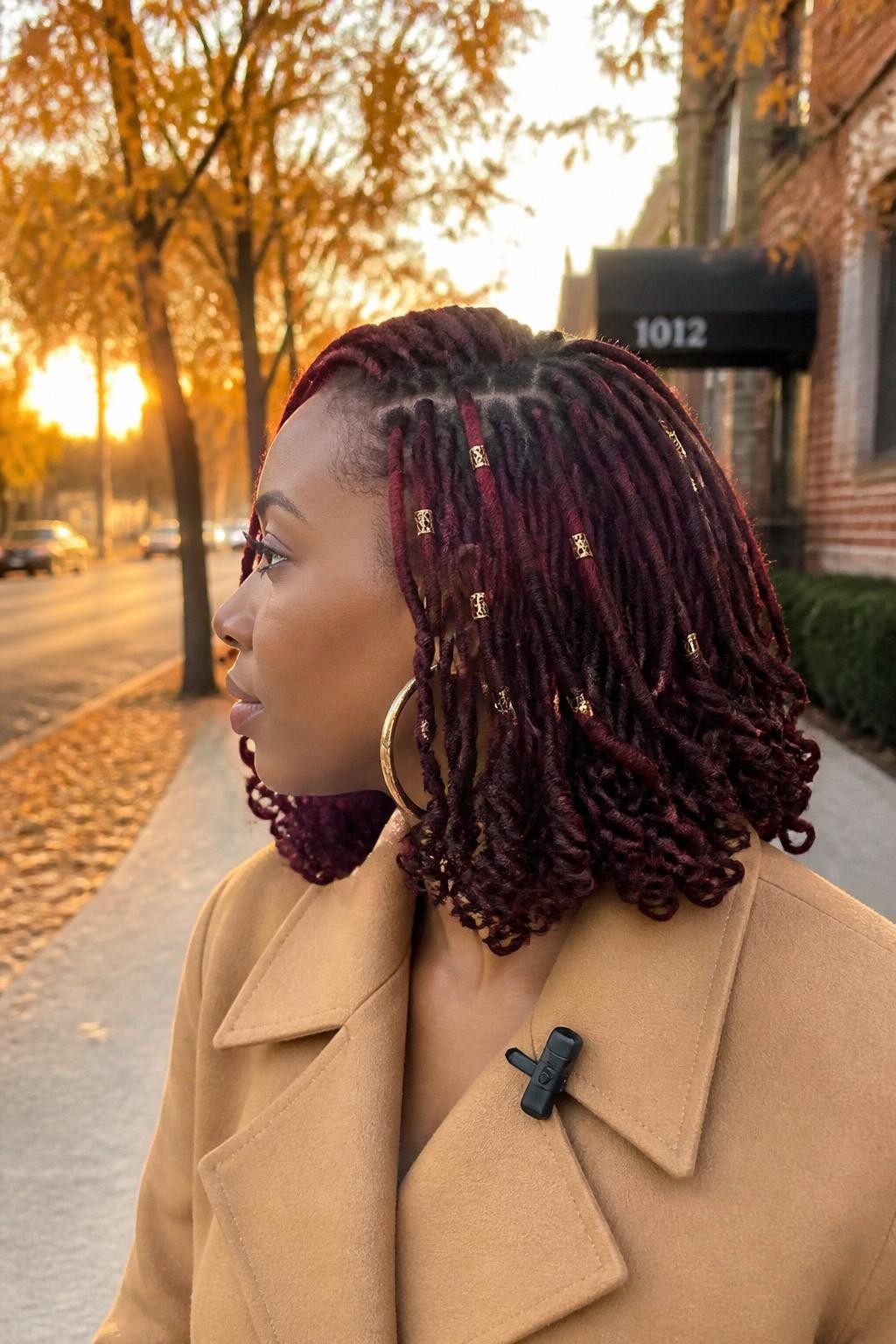 Photo of a Black woman wearing a shoulder-skimming curly loc lob in a deep burgundy-brown shade, with spiraled ends, a soft side part, and scattered copper cuffs around the front locs, side-profile view against a softly blurred fall street with amber trees, golden hour glow, large hoop earrings visible and a small clip attached to her coat collar, realistic loc texture and candid movement, latest iPhone photo quality.