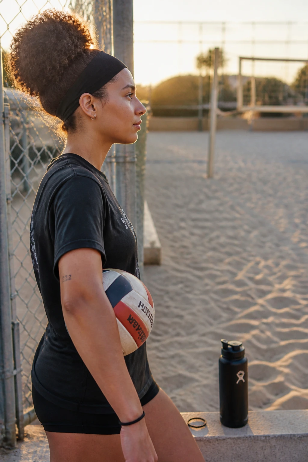 Image prompt

Photo of a woman wearing a high rounded curly puff held back with a wide moisture-wicking sports headband, side profile view beside a softly blurred outdoor volleyball court, golden hour glow, containing a small lived-in detail such as a volleyball tucked beneath her arm, a sports bottle near the fence, and a spare hair tie on her wrist, realistic candid pre-game photograph, latest iPhone photo quality.