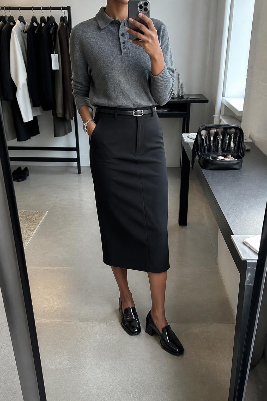 mirror selfie of a Black woman wearing a gray fine-knit polo sweater, black structured midi skirt, black leather belt, black heeled loafers, and silver watch, clearly showing the same clothing items, colors, fabrics, and accessories listed in the Outfit Pieces section, holding a phone in front of a mirror, cropped at NECK so the face does not show, with a relaxed casual selfie pose, in a fashion studio mirror corner, natural window light, including a clothing rack with garments behind and a makeup brush or cosmetic bag on the counter, latest iPhone photo quality.