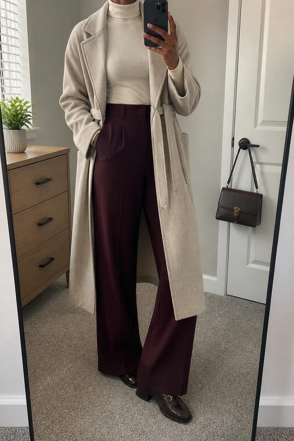 mirror selfie of a Black woman wearing an oatmeal wool wrap coat, fitted cream turtleneck, burgundy wide-leg trousers, brown heeled loafers, and a dark brown structured handbag, clearly showing the same clothing items, colors, fabrics, and accessories listed in the Outfit Pieces section, holding a phone in front of a mirror, cropped at NECK so the face does not show, with a relaxed casual selfie pose, in a minimal dressing room, natural window light, including a small plant on a dresser and a handbag hanging on a door handle, latest iPhone photo quality.