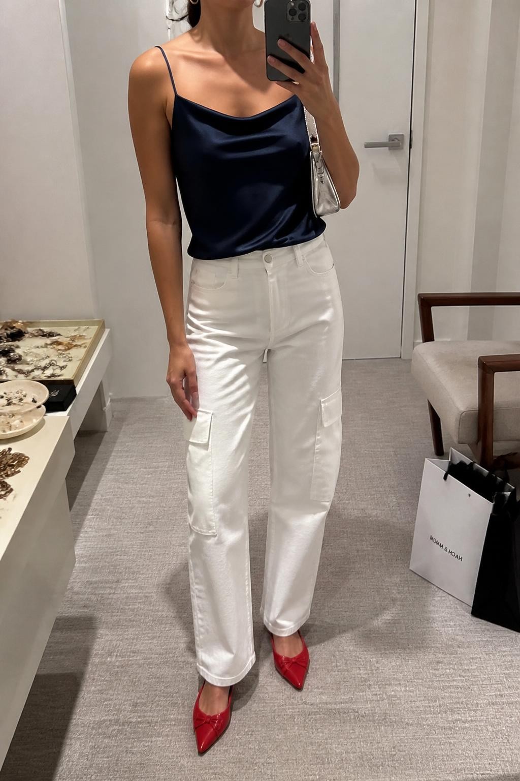 mirror selfie of a woman wearing a navy satin camisole tucked into white relaxed straight-leg cargo pants, red pointed slingback flats, silver mini shoulder bag, and delicate silver hoop earrings, clearly showing the same clothing items, colors, fabrics, and accessories listed in the Outfit Pieces section, holding a phone in front of a mirror, cropped at NECK so the face does not show, with a relaxed casual selfie pose, in a minimal dressing room, soft indoor lighting, including a jewelry tray on the vanity and shopping bags beside a chair, latest iPhone photo quality.