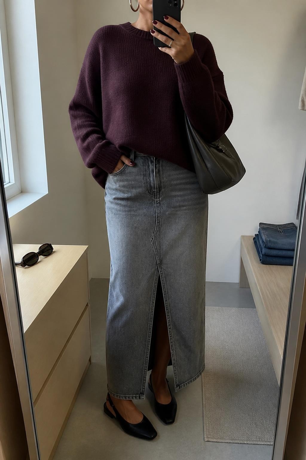 mirror selfie of a woman wearing the exact outfit described above, clearly showing a gray denim maxi skirt with a front slit, plum oversized knit sweater, black slingback flats, a charcoal shoulder bag, and silver hoop earrings, holding a phone in front of a mirror, cropped at NECK so the face does not show, with a relaxed casual selfie pose, in a minimal dressing room, natural window light, with sunglasses placed on a dresser and folded denim jeans on a bench, latest iPhone photo quality.