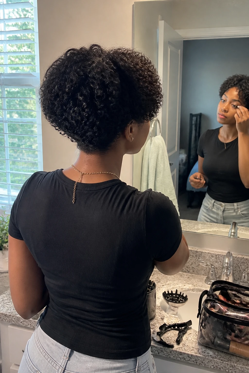 Image prompt

Photo of a Black woman wearing a stacked short curly bob with a softly lifted back, slightly longer curls around the front, and defined natural texture, back three-quarter view highlighting the rounded stacked shape, standing at a bathroom vanity mirror, natural window light, containing a small lived-in detail such as sectioning clips on the counter, a diffuser nearby, and a makeup bag left open, realistic getting-ready moment, latest iPhone photo quality.