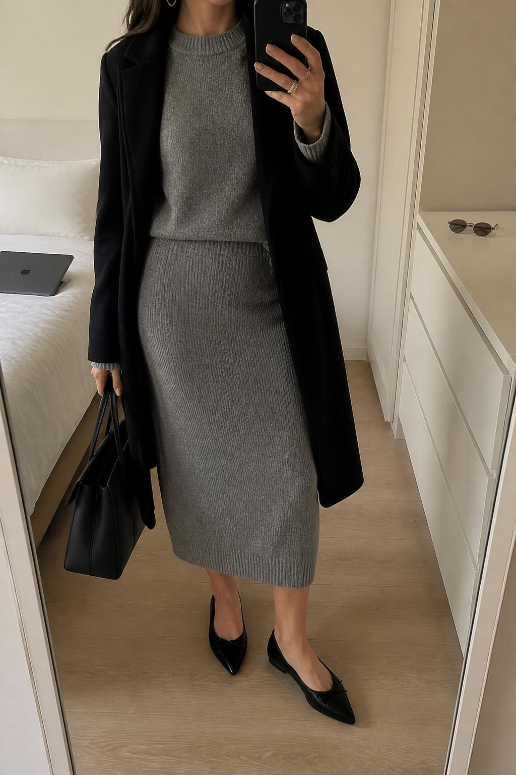 mirror selfie of a woman wearing the exact outfit described above, clearly showing a gray knit sweater, matching gray knit midi skirt, black wool coat worn open, black pointed flats, a structured black tote bag, and silver hoop earrings, holding a phone in front of a mirror, cropped at NECK so the face does not show, with a relaxed casual selfie pose, in a minimal dressing room, soft indoor lighting, with a laptop or tablet on the bed and sunglasses placed on a dresser, latest iPhone photo quality.