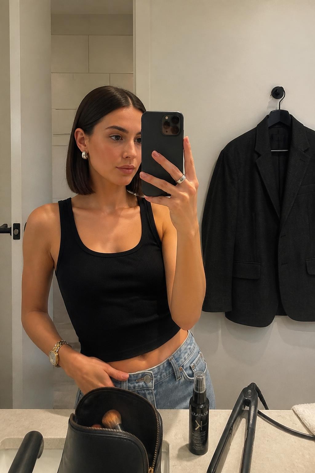 Photo of a woman wearing a glossy collarbone-length glass lob with a precise center part, straight smooth lengths, softly curved ends, and both sides tucked behind the ears, candid mirror selfie at a modern bathroom vanity, soft indoor lighting, flat iron and smoothing serum beside an open makeup bag, sculptural earrings visible and a charcoal blazer hanging nearby, latest iPhone photo quality.