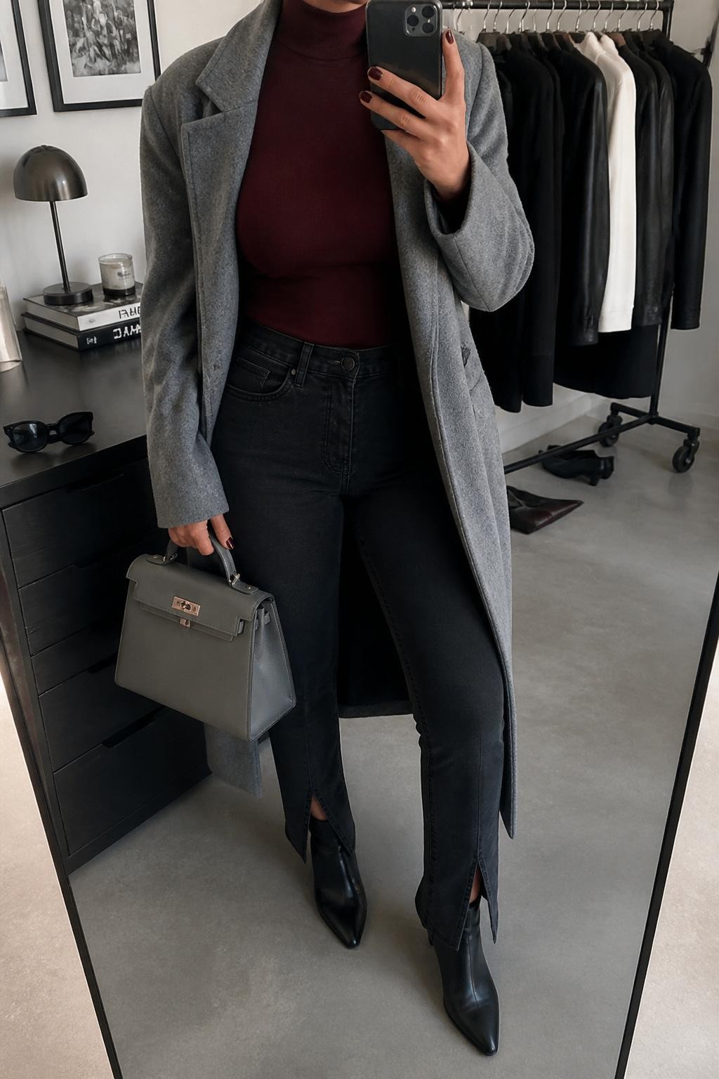 mirror selfie of a curvy woman wearing a gray wool coat over a burgundy fitted mock-neck top, black split-hem jeans, black pointed ankle boots, and a gray structured handbag, clearly showing the same clothing items, colors, fabrics, and accessories listed in the Outfit Pieces section, holding a phone in front of a mirror, cropped at NECK so the face does not show, with a relaxed casual selfie pose, in a fashion studio mirror corner, soft indoor lighting, including a clothing rack with garments behind and sunglasses placed on a dresser, latest iPhone photo quality.