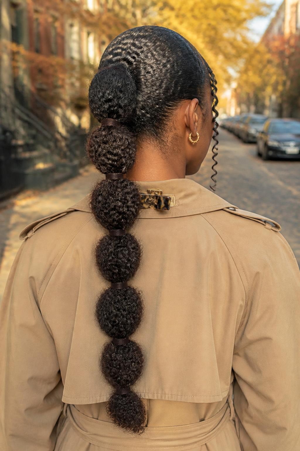 Photo of a Black woman wearing a long textured bubble ponytail with rounded curly sections separated by narrow chocolate velvet ties, softly laid edges, and two spiral tendrils framing her face, back-of-head view against a softly blurred fall street, golden hour lighting, hair clip attached to her trench coat collar and small gold earrings visible, realistic natural curl texture, latest iPhone photo quality.