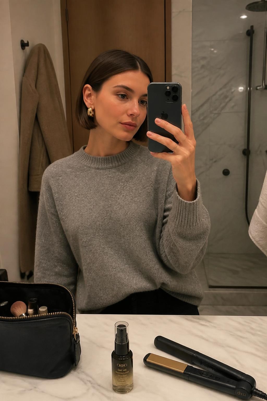 Photo of a woman wearing a smooth ear-length bob with a precise center part, softly curved ends, and both sides tucked neatly behind the ears, candid mirror selfie at a modern bathroom vanity, warm soft indoor lighting, flat iron and smoothing serum beside an open makeup pouch, sculptural gold earrings visible and a wool coat hanging in the background, latest iPhone photo quality.