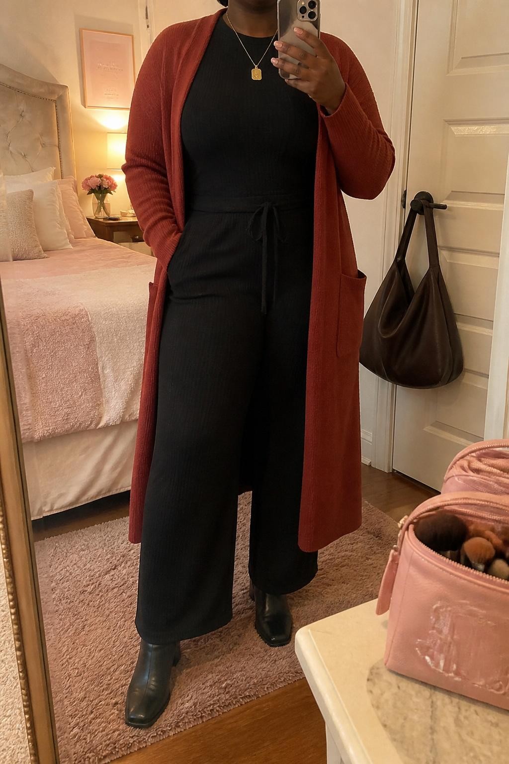 mirror selfie of a plus size Black woman wearing the exact outfit described above, a brick red longline coatigan layered over a black wide-leg knit jumpsuit, black leather block heel ankle boots, brushed gold pendant necklace, and dark brown soft hobo bag, clearly showing the same clothing items, colors, fabrics, and accessories listed in the Outfit Pieces section, holding a phone in front of a mirror, cropped at NECK so the face does not show, with a relaxed casual selfie pose, in a feminine-themed bedroom, evening golden hour, including a handbag hanging on a door handle and a makeup brush or cosmetic bag on the counter, latest iPhone photo quality.