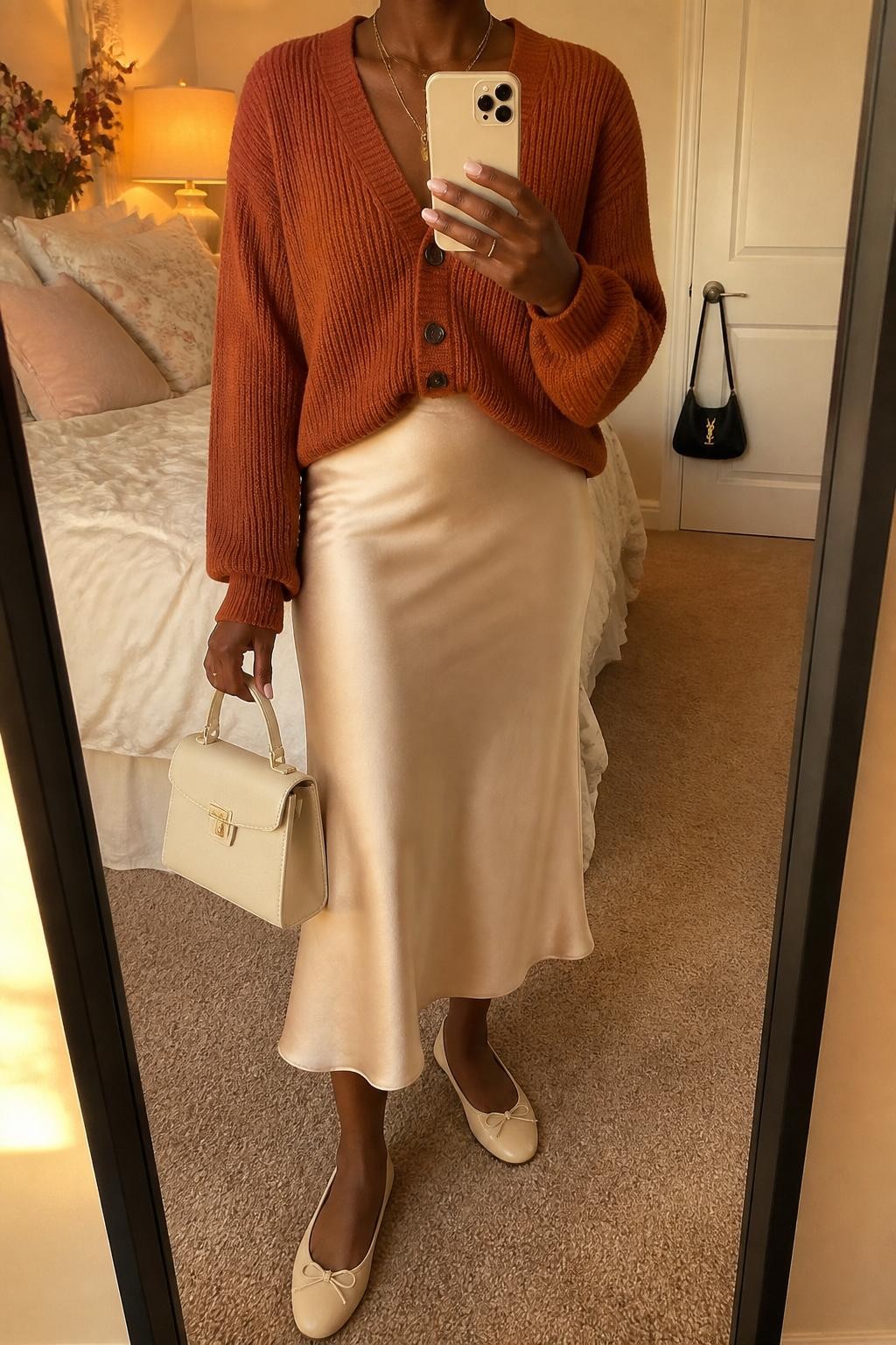 Mirror selfie of a Black woman with rich dark skin wearing a chunky rust-orange cardigan tucked slightly into a champagne satin midi skirt, cream leather ballet flats, a small cream top-handle bag, and layered delicate gold necklaces, clearly showing the exact clothing items, colors, textures, and accessories listed, holding a latest-model iPhone in front of a mirror, cropped at the neck so the face does not show, relaxed casual selfie pose, in a feminine-themed bedroom with evening golden-hour light, soft pillows on an unmade bed and a handbag hanging on a door handle, realistic uncluttered composition, latest iPhone photo quality.