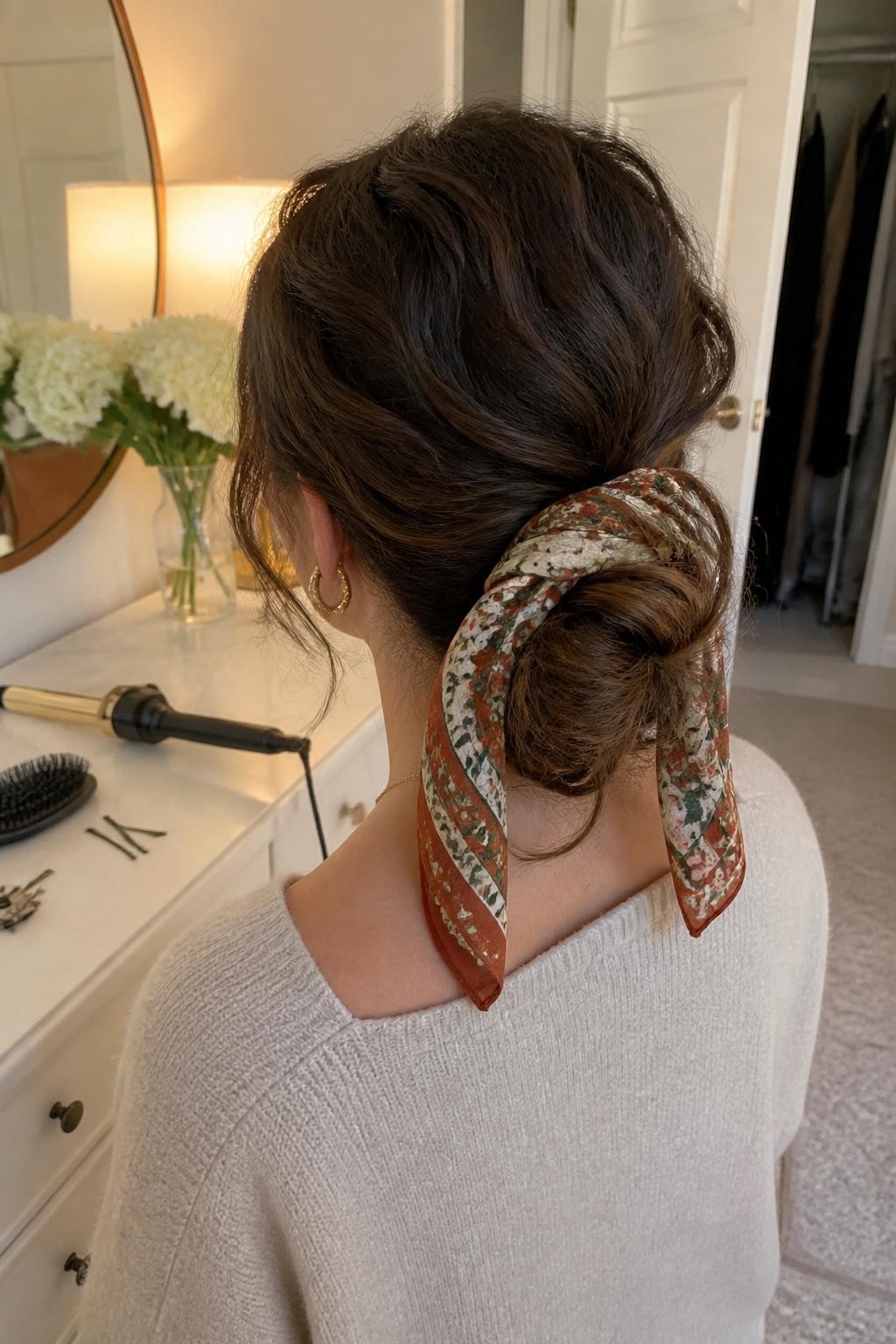 Photo of a woman wearing long curls arranged into a soft low bun with visible curled loops, gentle crown volume, and a narrow rust, olive, and cream printed silk scarf wrapped around the bun, back-of-head view in a warmly lit bedroom, curling wand and bobby pins on the dresser beside a hairbrush, gold earrings peeking through and a slightly open closet behind her, realistic loose tendrils and natural texture, latest iPhone photo quality.