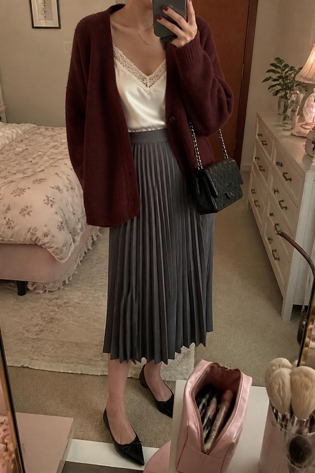 mirror selfie of a woman wearing a burgundy soft cardigan, white silk camisole, gray pleated midi skirt, black pointed flats, and black quilted shoulder bag, clearly showing the same clothing items, colors, fabrics, and accessories listed in the Outfit Pieces section, holding a phone in front of a mirror, cropped at NECK so the face does not show, with a relaxed casual selfie pose, in a feminine-themed bedroom, warm moody lighting, including a makeup brush or cosmetic bag on the counter and a small plant on a dresser, latest iPhone photo quality.