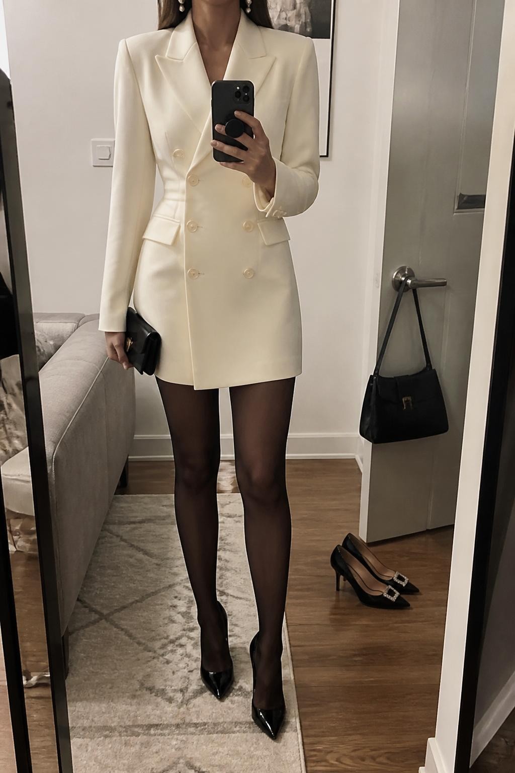 mirror selfie of a woman wearing the exact outfit described above, clearly showing an ivory blazer dress, sheer black tights, black pointed pumps, pearl earrings, and a small black clutch, holding a phone in front of a mirror, cropped at NECK so the face does not show, with a relaxed casual selfie pose, in a stylish city apartment, soft indoor lighting, with a pair of heels on the floor nearby and a handbag hanging on a door handle, latest iPhone photo quality.