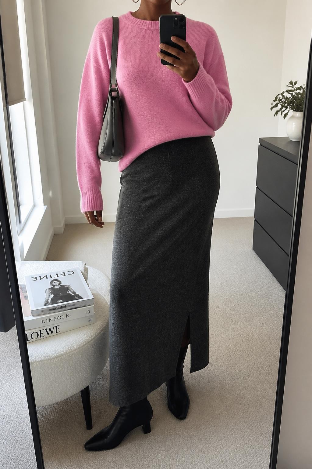 mirror selfie of a Black woman wearing a bubblegum pink crew-neck wool sweater, charcoal gray wool maxi skirt with a back slit, black leather ankle boots, silver hoop earrings, and a structured gray shoulder bag, clearly showing the same clothing items, colors, fabrics, and accessories listed in the Outfit Pieces section, holding a phone in front of a mirror, cropped at NECK so the face does not show, with a relaxed casual selfie pose, in a minimal dressing room, natural window light, including a stack of fashion magazines on a side table and a small plant on a dresser, latest iPhone photo quality.