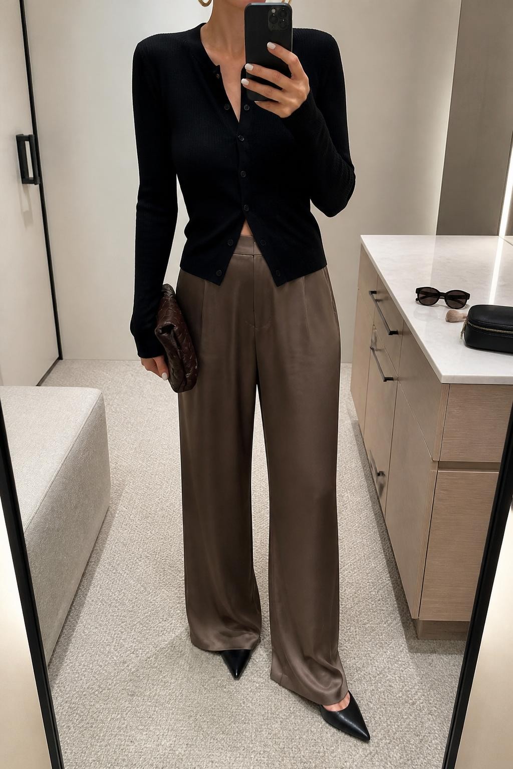 mirror selfie of a woman wearing the exact outfit described above, clearly showing mocha satin wide-leg pants, a fitted black cardigan buttoned at the center, black pointed mules, a dark brown clutch, and gold hoop earrings, holding a phone in front of a mirror, cropped at NECK so the face does not show, with a relaxed casual selfie pose, in a minimal dressing room, soft indoor lighting, with sunglasses placed on a dresser and a makeup brush or cosmetic bag on the counter, latest iPhone photo quality.