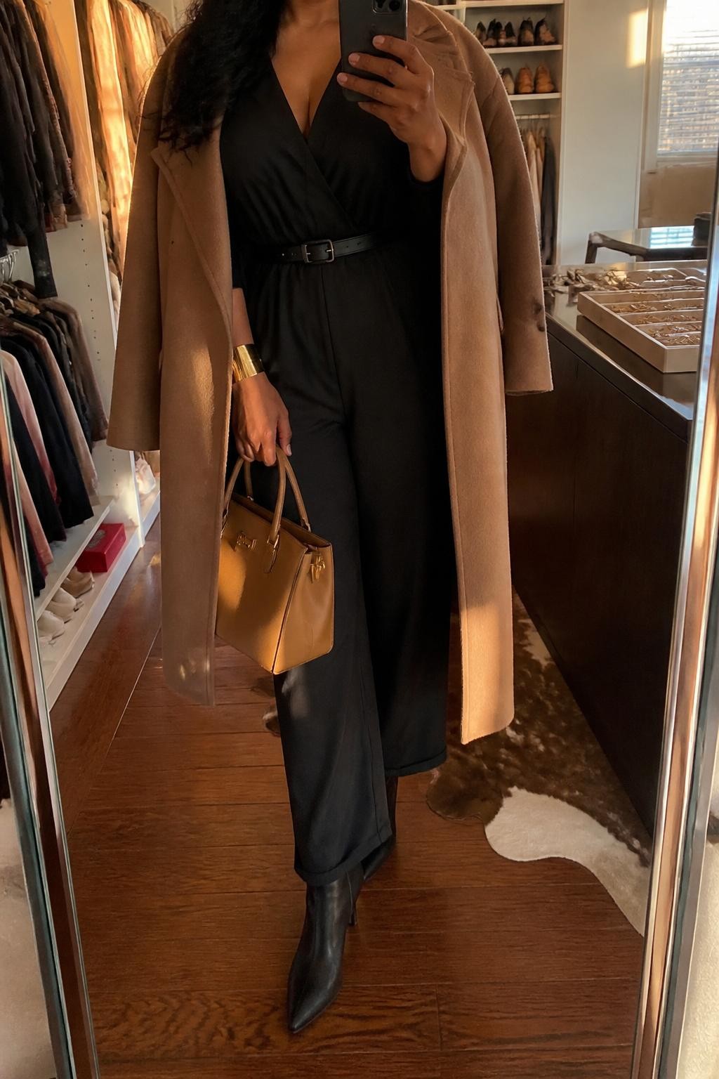 mirror selfie of an Afro-Latina woman wearing a black belted jumpsuit, camel wool coat, black heeled boots, gold cuff bracelet, and a camel structured handbag, holding a phone in front of a mirror, cropped at NECK so the face does not show, relaxed casual selfie pose, in a walk-in closet mirror with evening golden hour lighting, including open closet with clothes visible and a jewelry tray on the vanity, realistic latest iPhone photo quality.