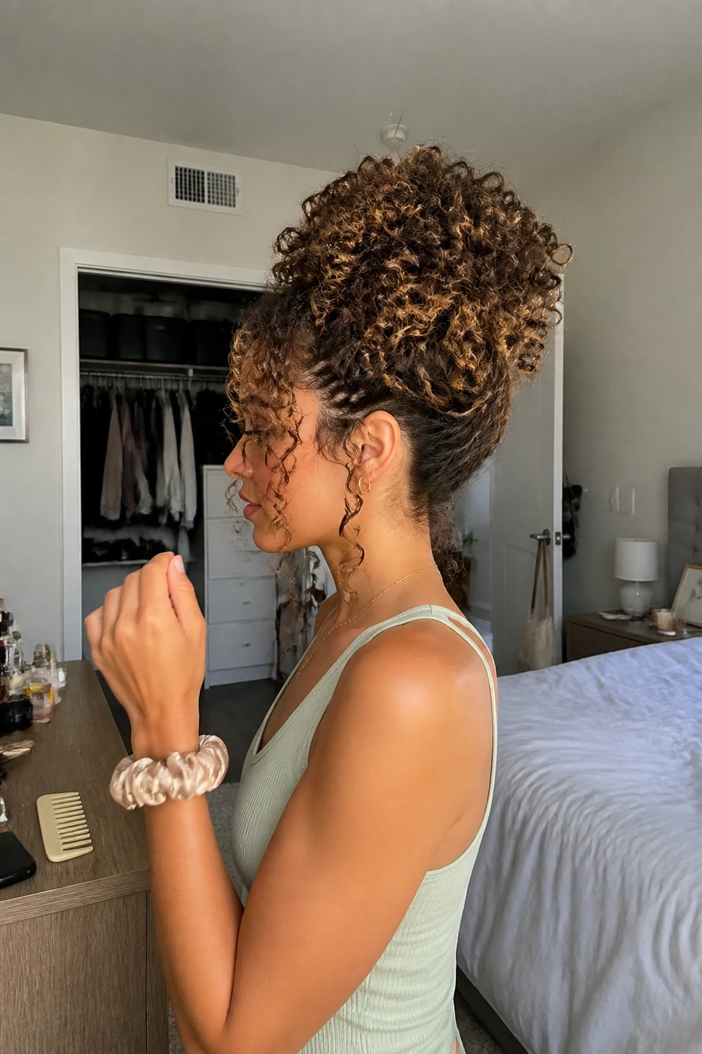 Image prompt

Photo of a woman wearing a soft high curly pineapple updo with full defined curls gathered loosely at the crown and two small face-framing tendrils, side profile view highlighting the volume and curl texture, standing in a sun-drenched bedroom with a slightly open closet behind her, natural window light, containing a small lived-in detail such as a silk scrunchie on her wrist and a wide-tooth comb on the dresser, realistic getting-ready moment, latest iPhone photo quality.