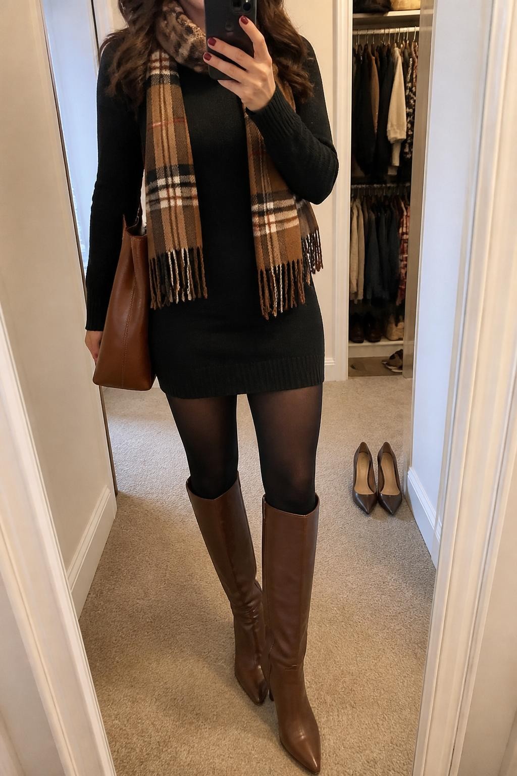 mirror selfie of a woman wearing a black long-sleeve sweater dress, brown plaid scarf, black opaque tights, brown knee-high boots, and a brown leather tote, clearly showing the same clothing items, colors, fabrics, and accessories listed in the Outfit Pieces section, holding a phone in front of a mirror, cropped at NECK so the face does not show, with a relaxed casual selfie pose, in a walk-in closet mirror, soft indoor lighting, including open closet with clothes visible and a pair of heels on the floor nearby, latest iPhone photo quality.
