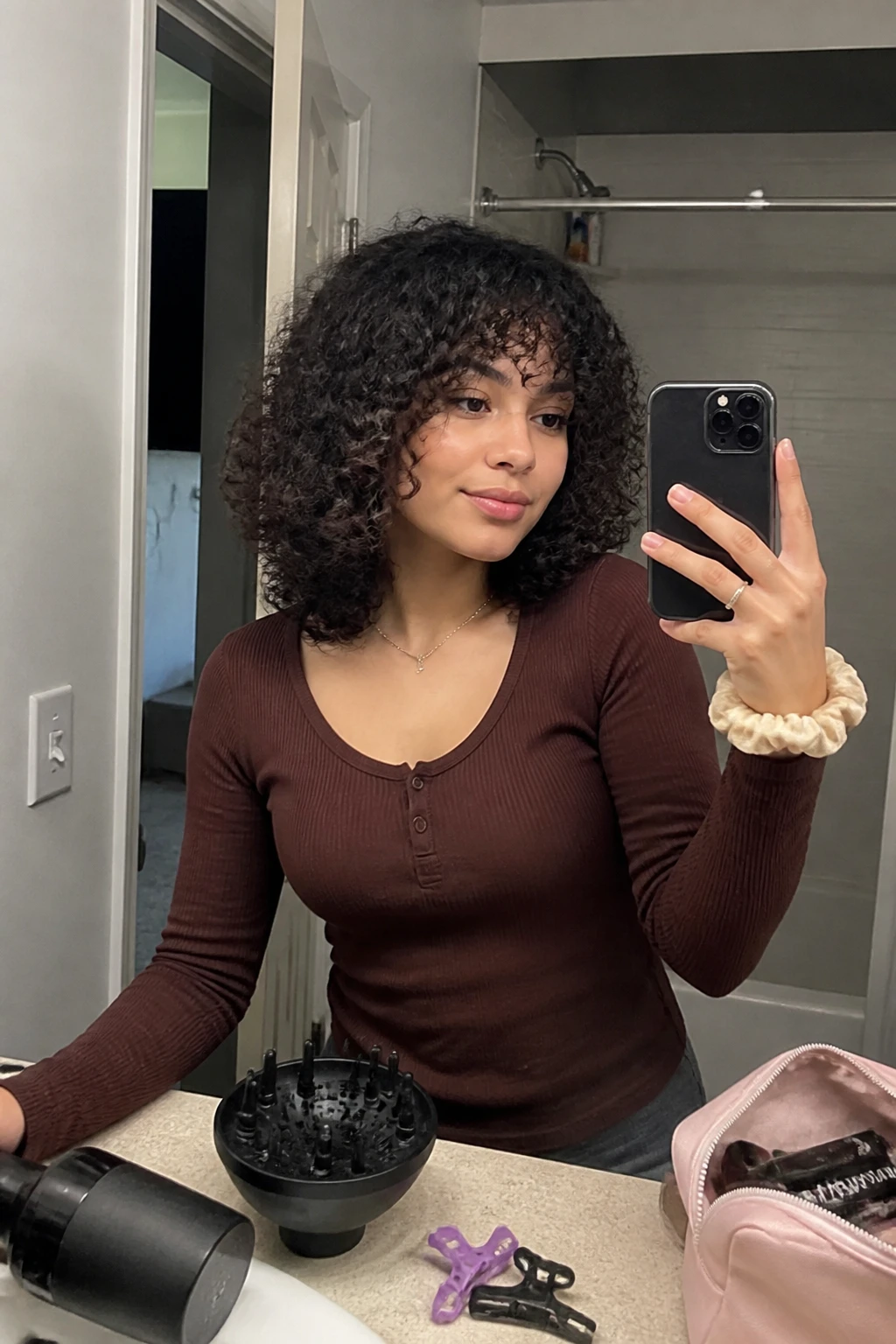 Image prompt
Photo of a woman wearing a medium-length 3B curly shag with short crown layers, soft curly bangs, and springy ringlets creating a light airy shape around the face, front three-quarter view in a bathroom mirror, soft indoor lighting, containing a small lived-in detail such as sectioning clips and a diffuser on the counter, a makeup bag nearby, and a silk scrunchie on her wrist, realistic getting-ready moment, latest iPhone photo quality