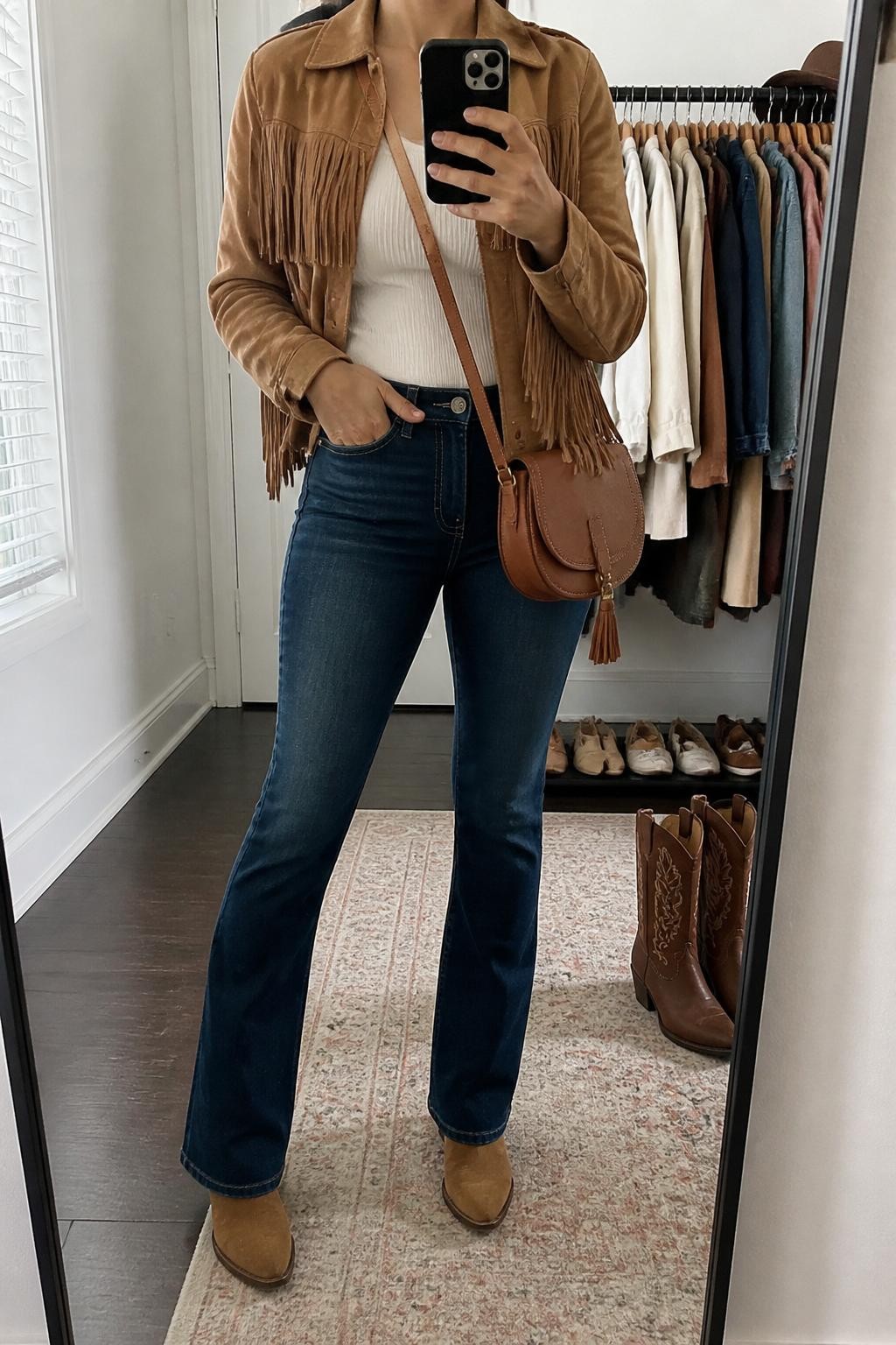 mirror selfie of a woman wearing the exact outfit described above, clearly showing a tan suede fringe jacket, fitted white ribbed top, dark blue bootcut jeans, brown western ankle boots, and a cognac crossbody saddle bag, holding a phone in front of a mirror, cropped at NECK so the face does not show, with a relaxed casual selfie pose, in a walk-in closet mirror, natural window light, with a clothing rack with garments behind and a pair of boots on the floor nearby, latest iPhone photo quality.